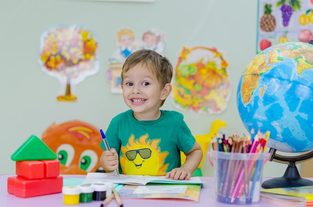 A smiling young boy sitting at a colorful desk with school supplies, including a globe, crayons, building blocks, and an open notebook, in a classroom decorated with educational posters and drawings.