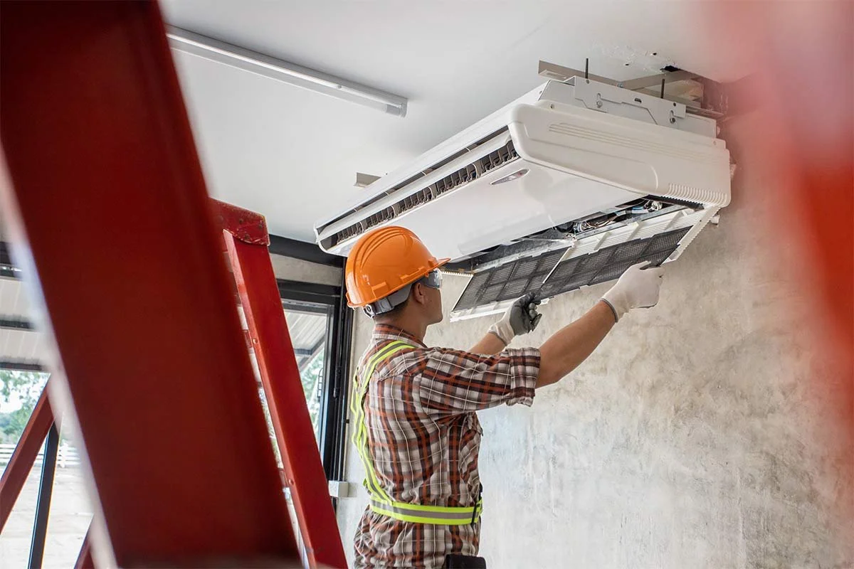 A worker installing or repairing a wall-mounted air conditioning unit, wearing an orange safety helmet, safety glasses, gloves, and a plaid shirt, in a room with a ladder nearby.