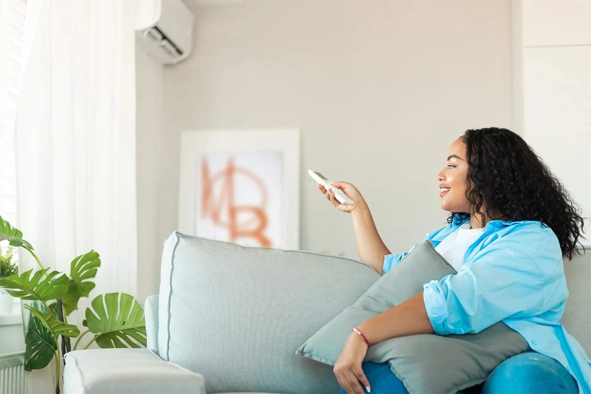 A woman with curly black hair smiling while sitting on a light-colored couch, holding a remote control and a pillow in a bright air conditioned living room with a plant and artwork in the background.