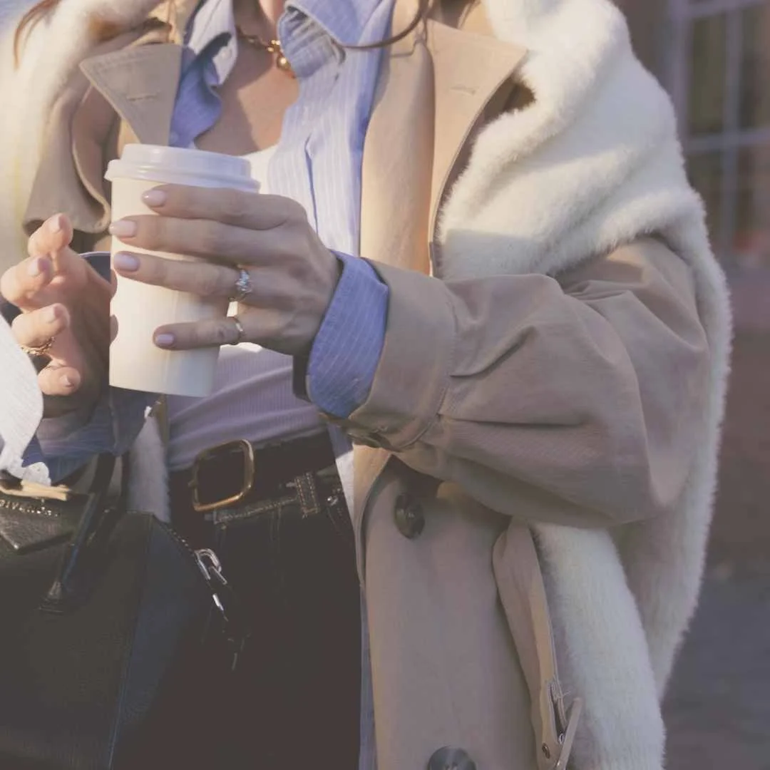 Person holding a paper coffee cup, wearing a beige coat over a blue shirt, with a ring on their finger and a handbag hanging from their shoulder.