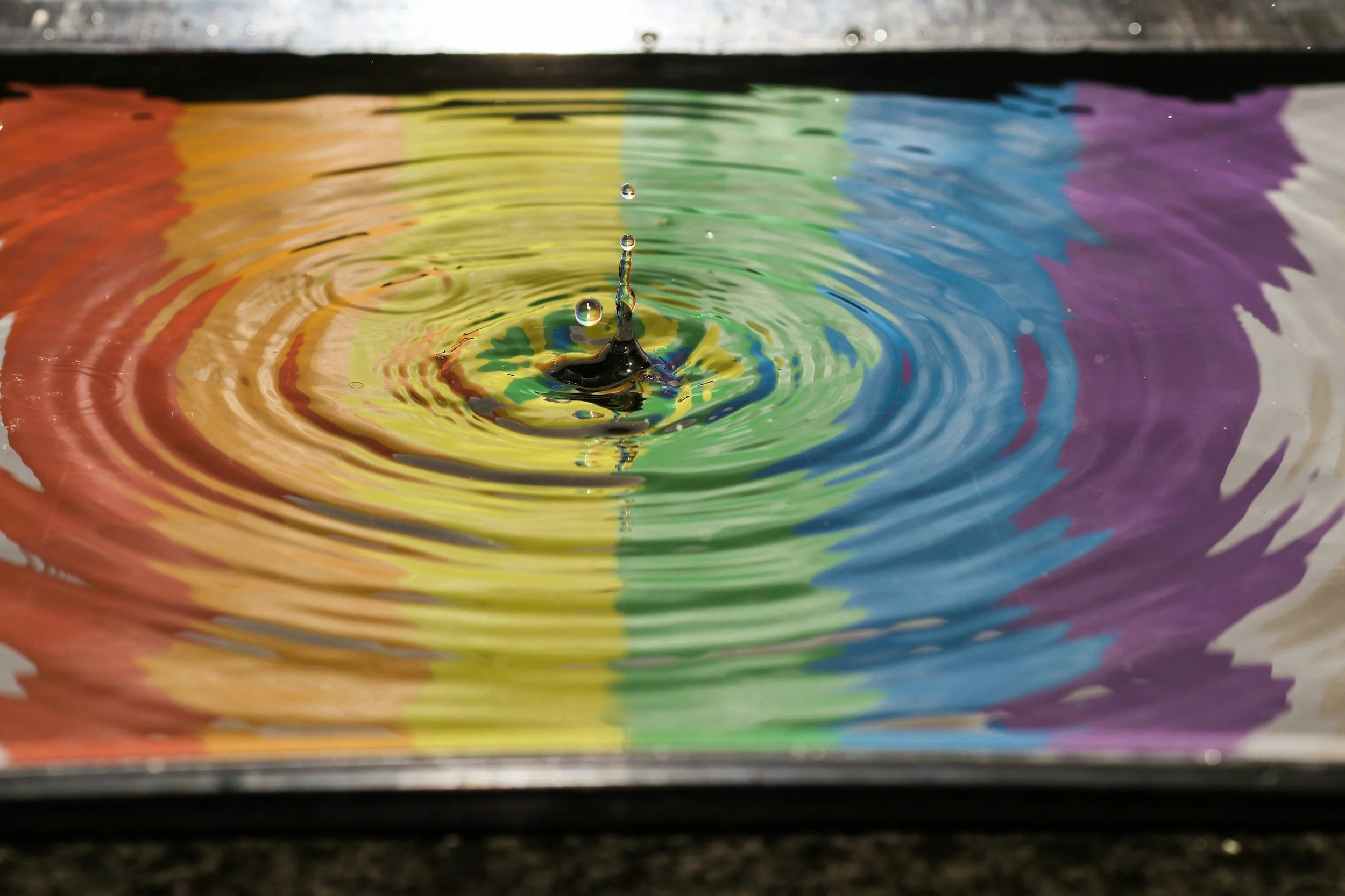 A close-up photo of a water droplet causing ripples in a multicolored liquid surface, with rainbow-like colors reflecting on the water.