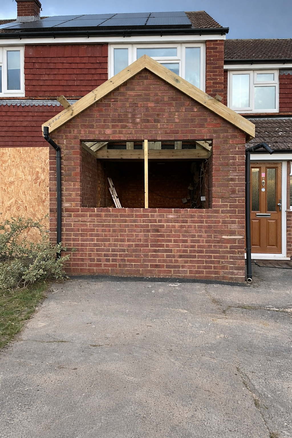 A house undergoing construction with a brick facade and a new front wall being built. The upper part of the house has solar panels and white-framed windows. The house has black gutters, and the area in front is paved with asphalt. A wooden frame structure is being built for the new extension, with a ladder visible inside.