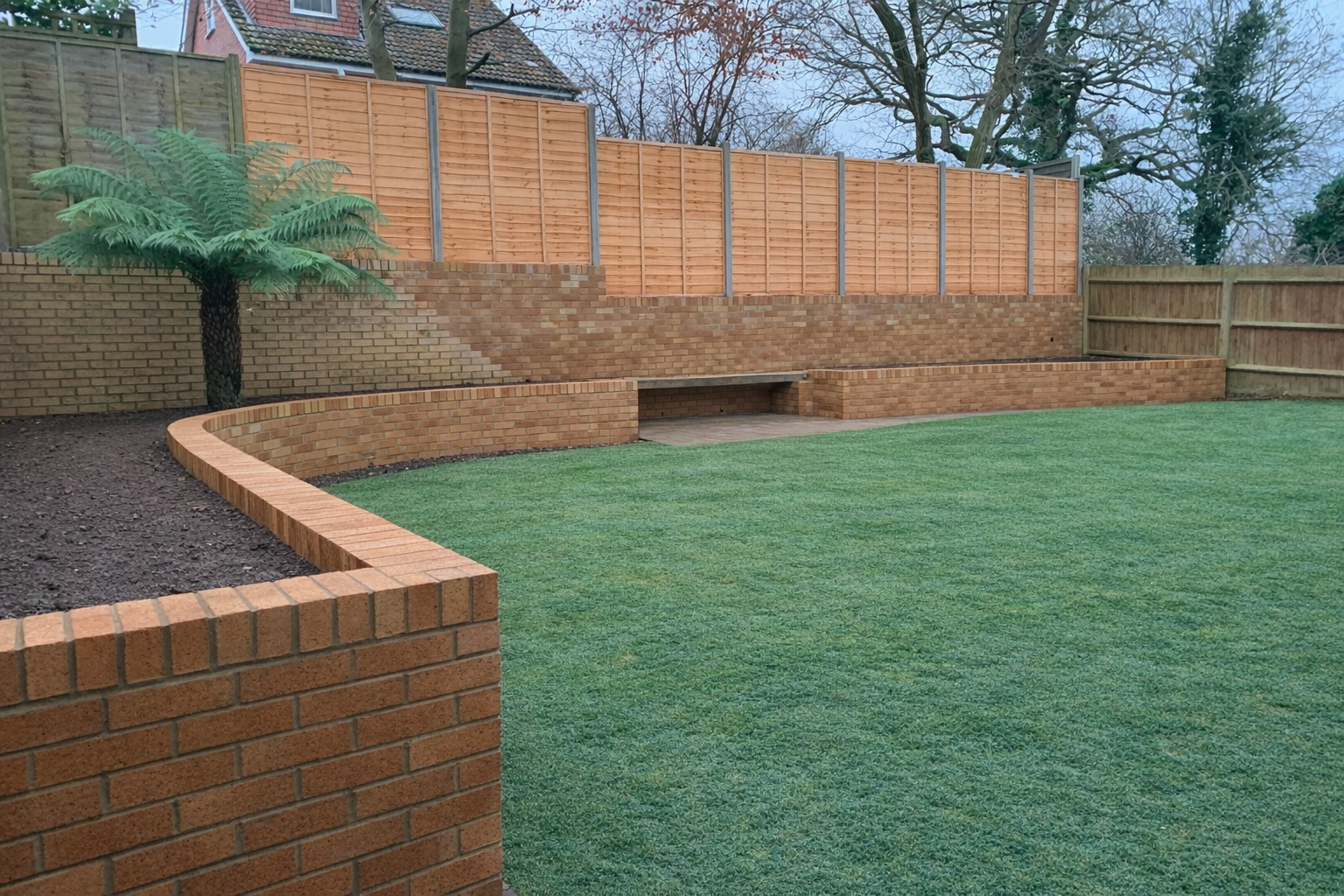 Backyard with artificial grass lawn, brick retaining wall, and wooden fencing, with a tree visible in the yard.