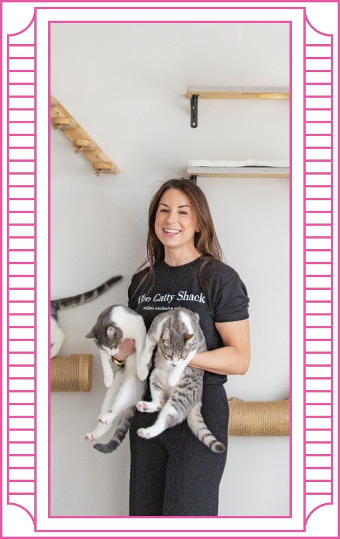 A woman smiling and holding two cats in an indoor room with cat shelves on the wall. The woman is wearing a black t-shirt with the words "The Catty Shack" on it. There is a pink border around the image.
