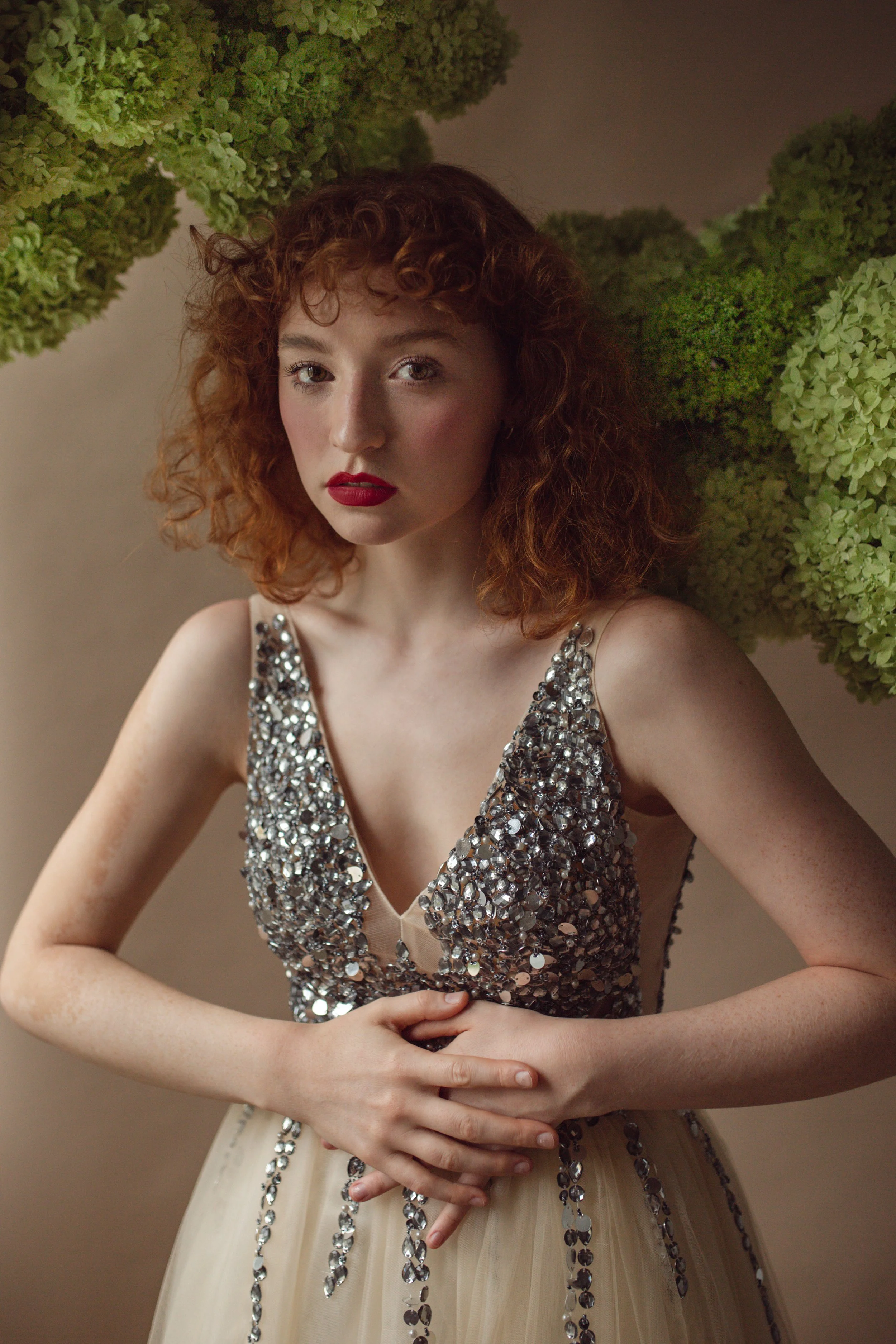 A young woman with curly red hair and makeup featuring red lipstick, wearing a sleeveless dress adorned with silver sequins, standing against a background of green hydrangea flowers.