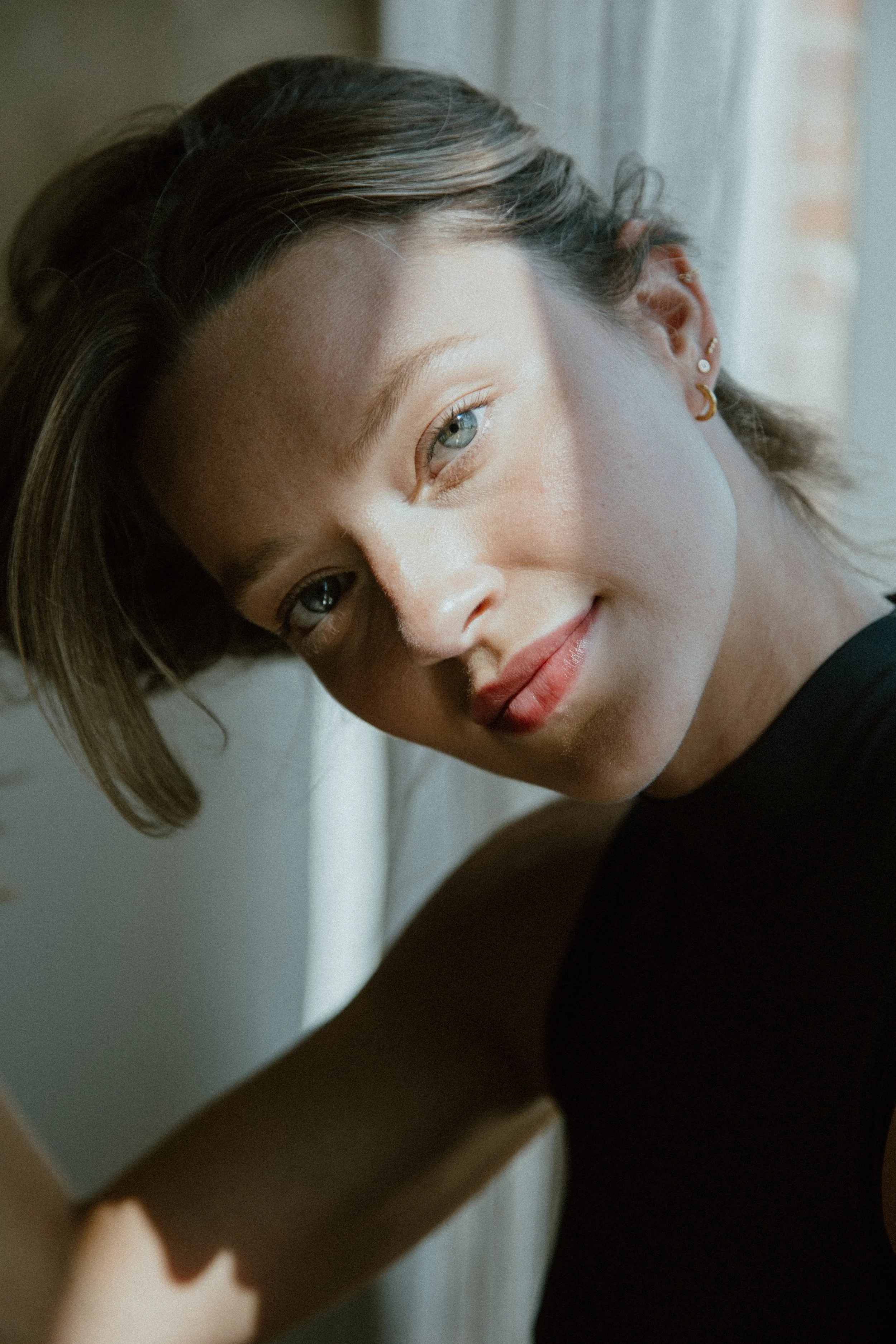 A woman with short brown hair and blue eyes smiling softly, with sunlight illuminating her face, sitting near a window.