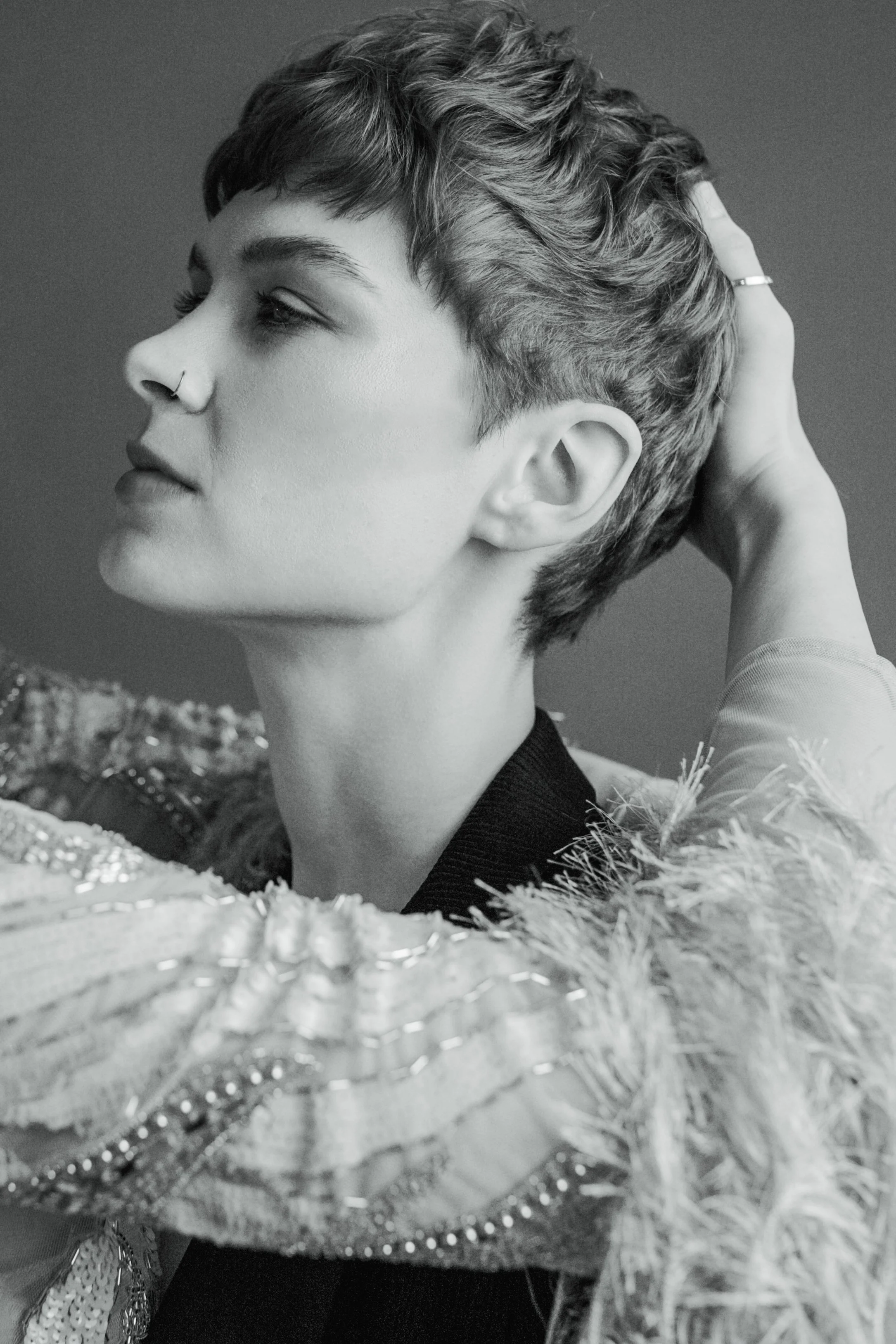 Black and white portrait of a woman with short, styled hair, wearing a nose ring and a feathered, embellished garment, resting her hand on the back of her head.