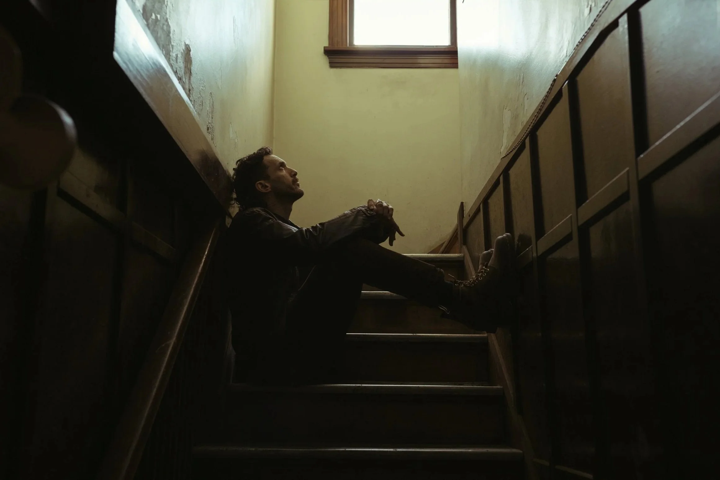 A person sitting on a staircase at the corner of a dimly lit interior space, gazing up towards a window at the top of the stairs, with worn yellow walls and wooden trim.