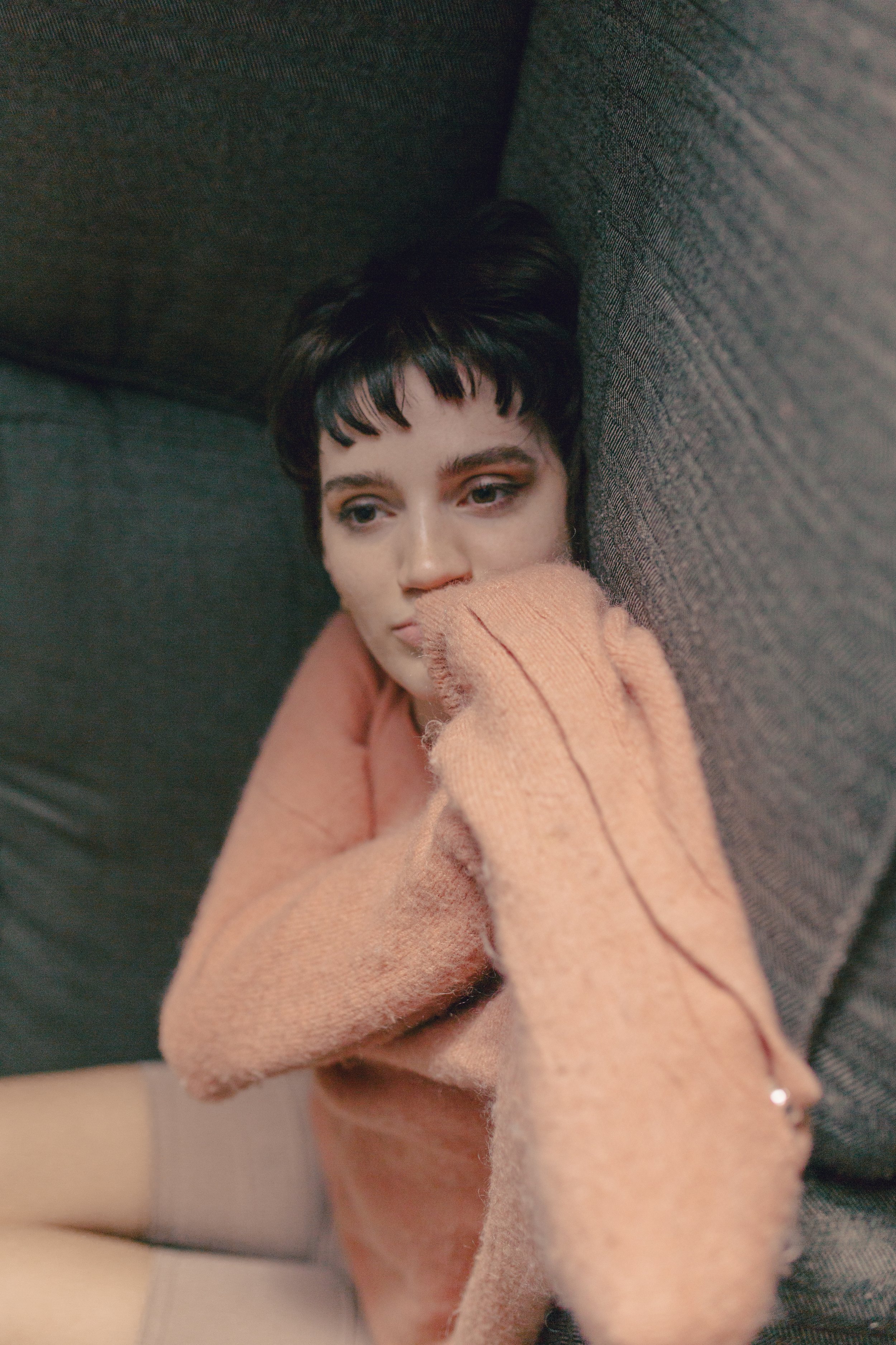 A young woman with dark short hair and bangs, wearing a peach-colored sweater, sitting against a dark gray fabric surface, with her face resting on her arm and looking contemplative.