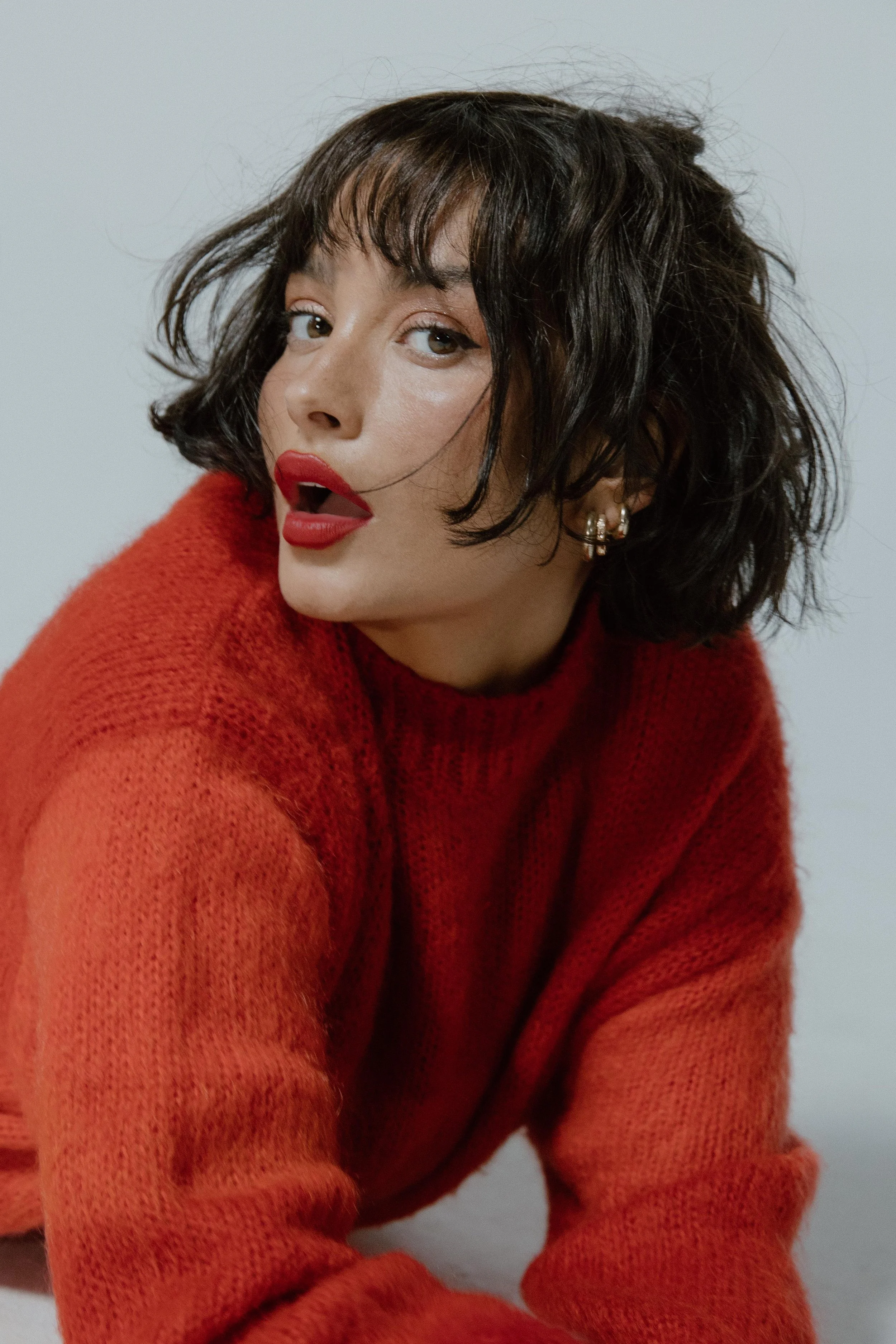 A woman with short, dark, wavy hair and red lipstick wearing a red, fuzzy sweater posing with her mouth slightly open.