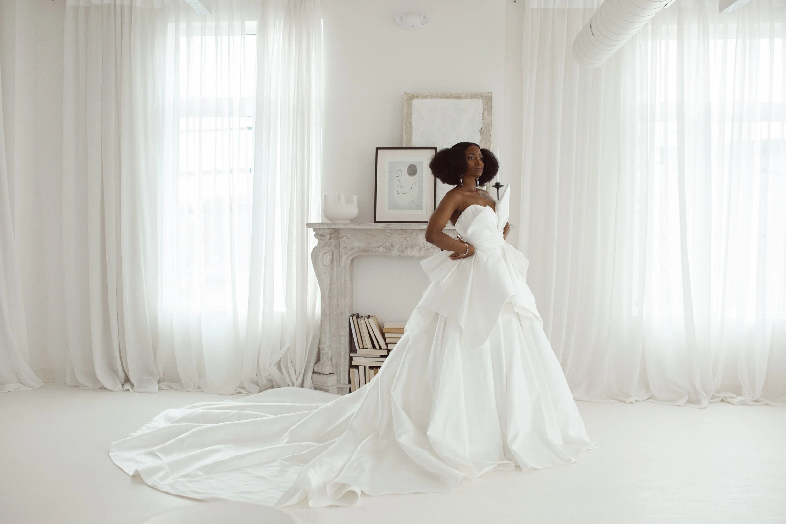 A woman in a white wedding gown standing in a bright white room with large windows and sheer curtains.