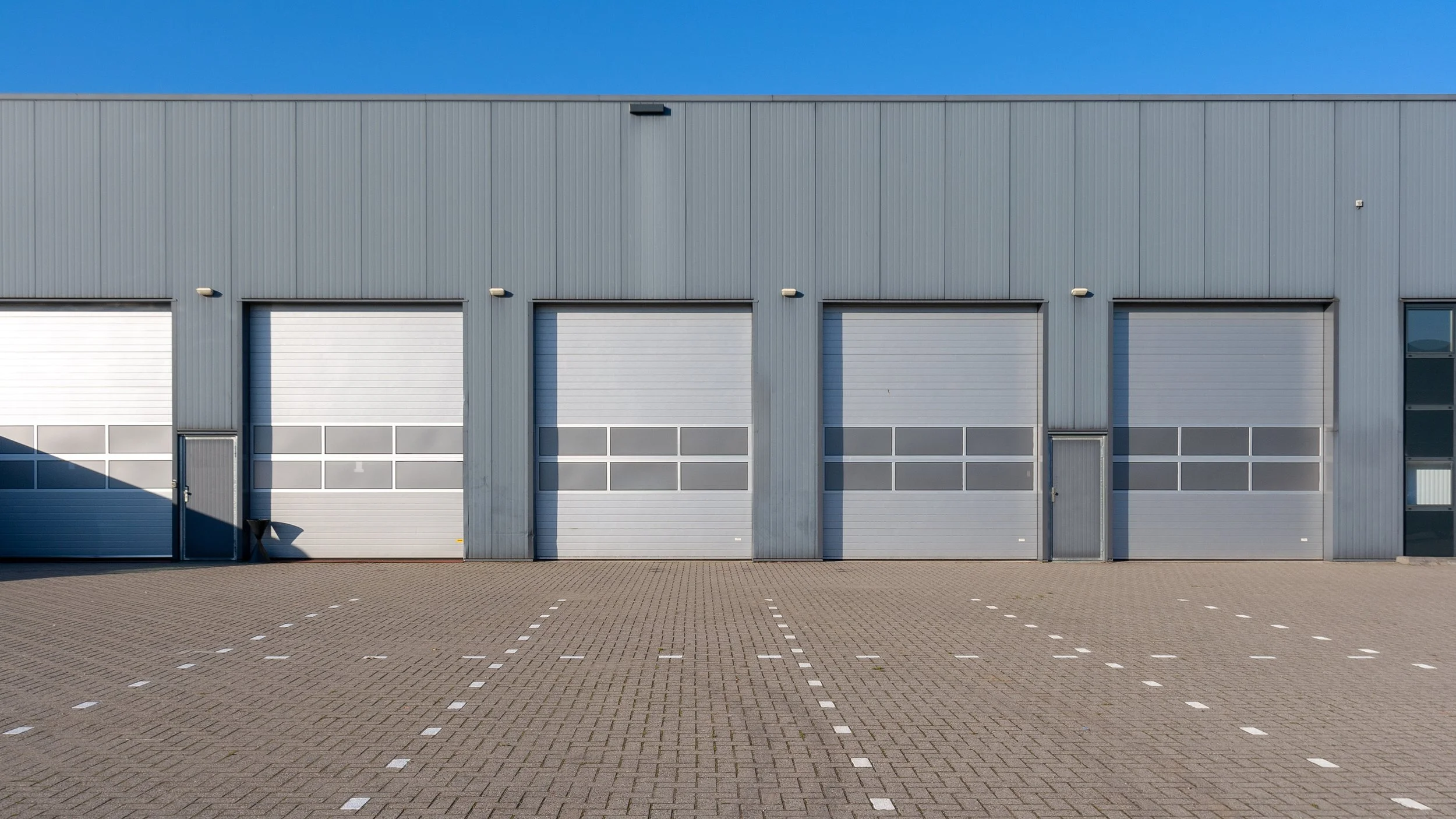 Front view of a large industrial warehouse with four garage doors, two small doors, and a paved parking lot in front.