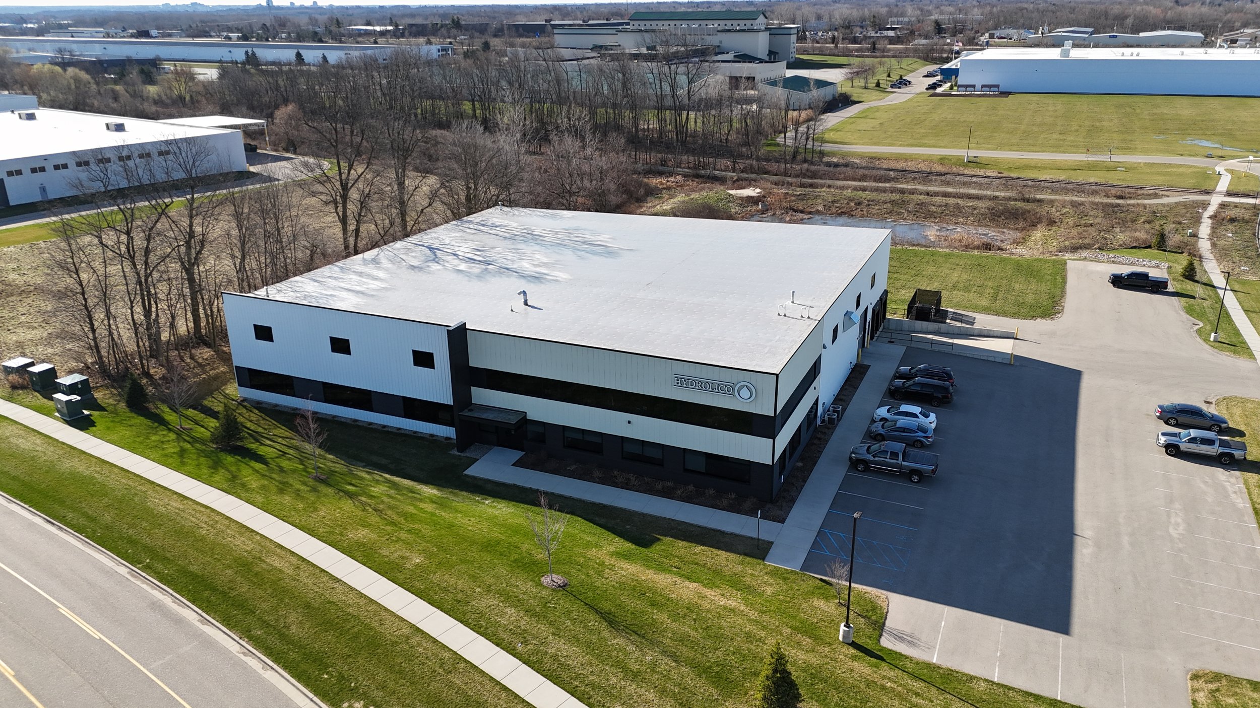 An aerial view of a commercial building with a parking lot, surrounded by grass and trees, in a business park or industrial area.