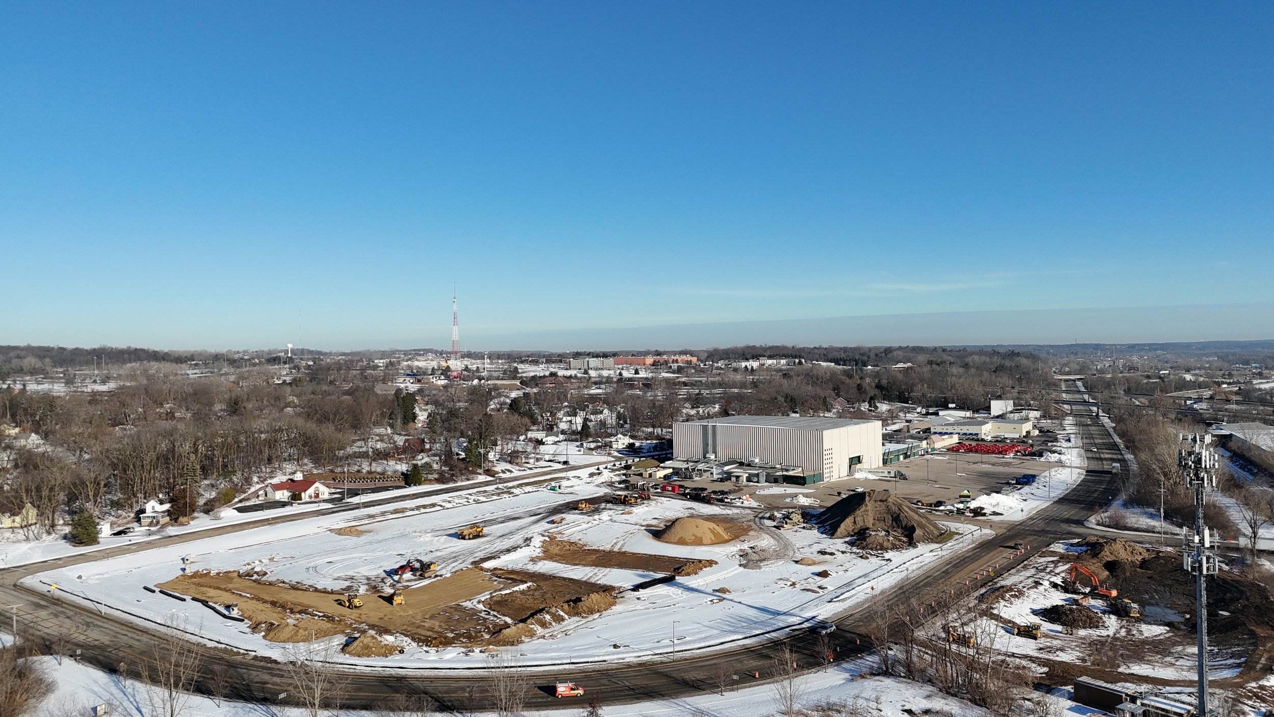 Construction site in a snowy landscape with machinery and piles of dirt, near a large industrial building and a railway track under a clear blue sky.
