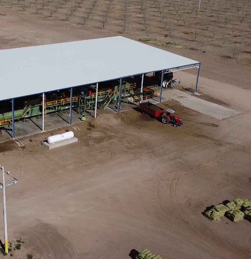 An aerial view of an agricultural facility with a large metal shed, machinery inside, a red tractor, and rows of hay bales outside on dry land.