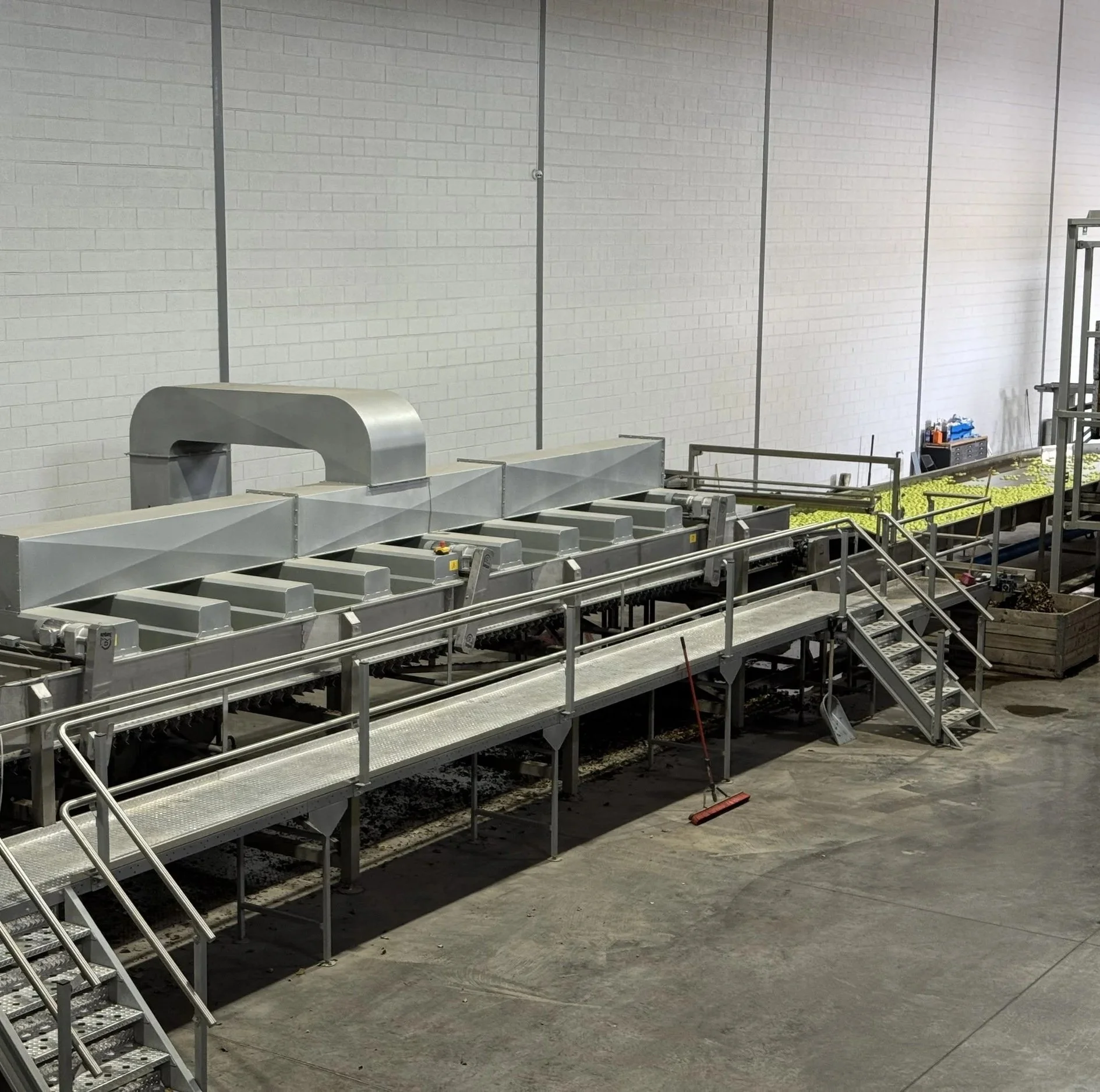 Industrial food processing conveyor system with metal rollers and stairs, inside a facility with white brick walls.