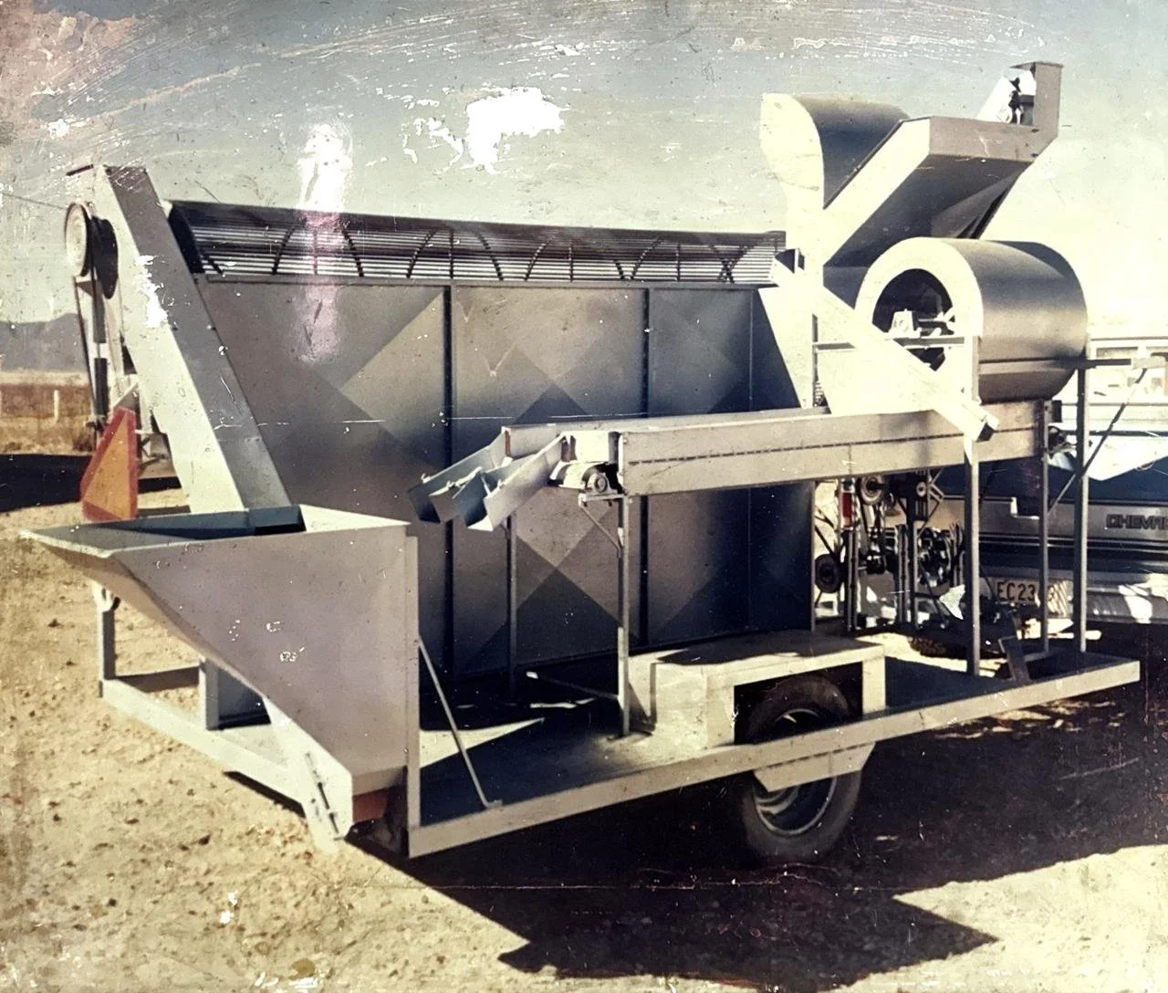 A large piece of industrial machinery or equipment mounted on a trailer, with several metal components, wheels, and belts, situated outdoors on a dirt surface.