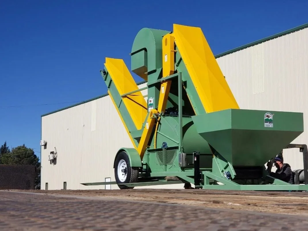 Green and yellow agricultural machine with a hopper setup outside near a beige building.