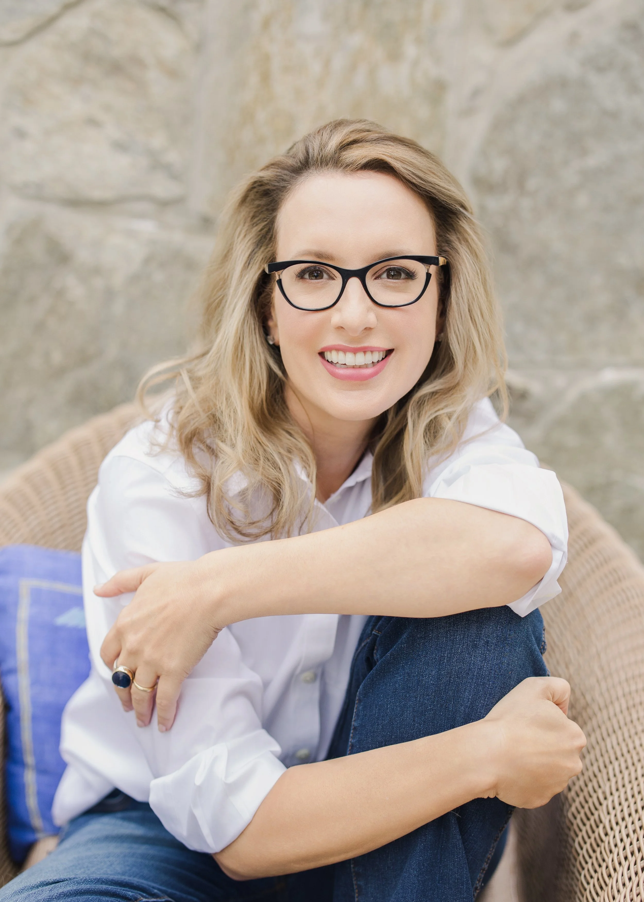 A smiling woman wearing glasses, a white shirt, and jeans, sitting on a wicker chair outdoors against a stone wall.