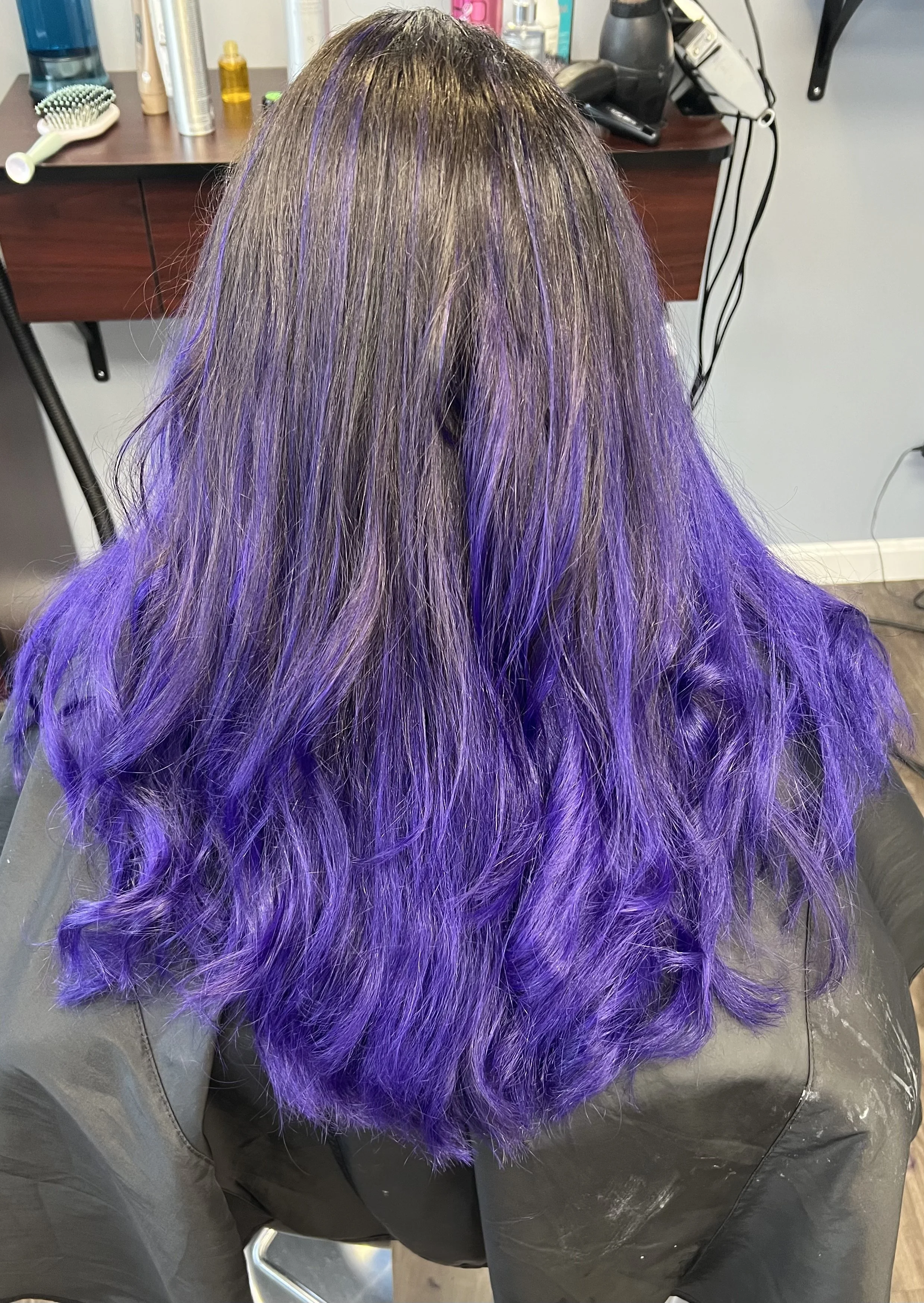 Back view of a person with long, wavy hair that is partially purple and grey, sitting in a salon chair.