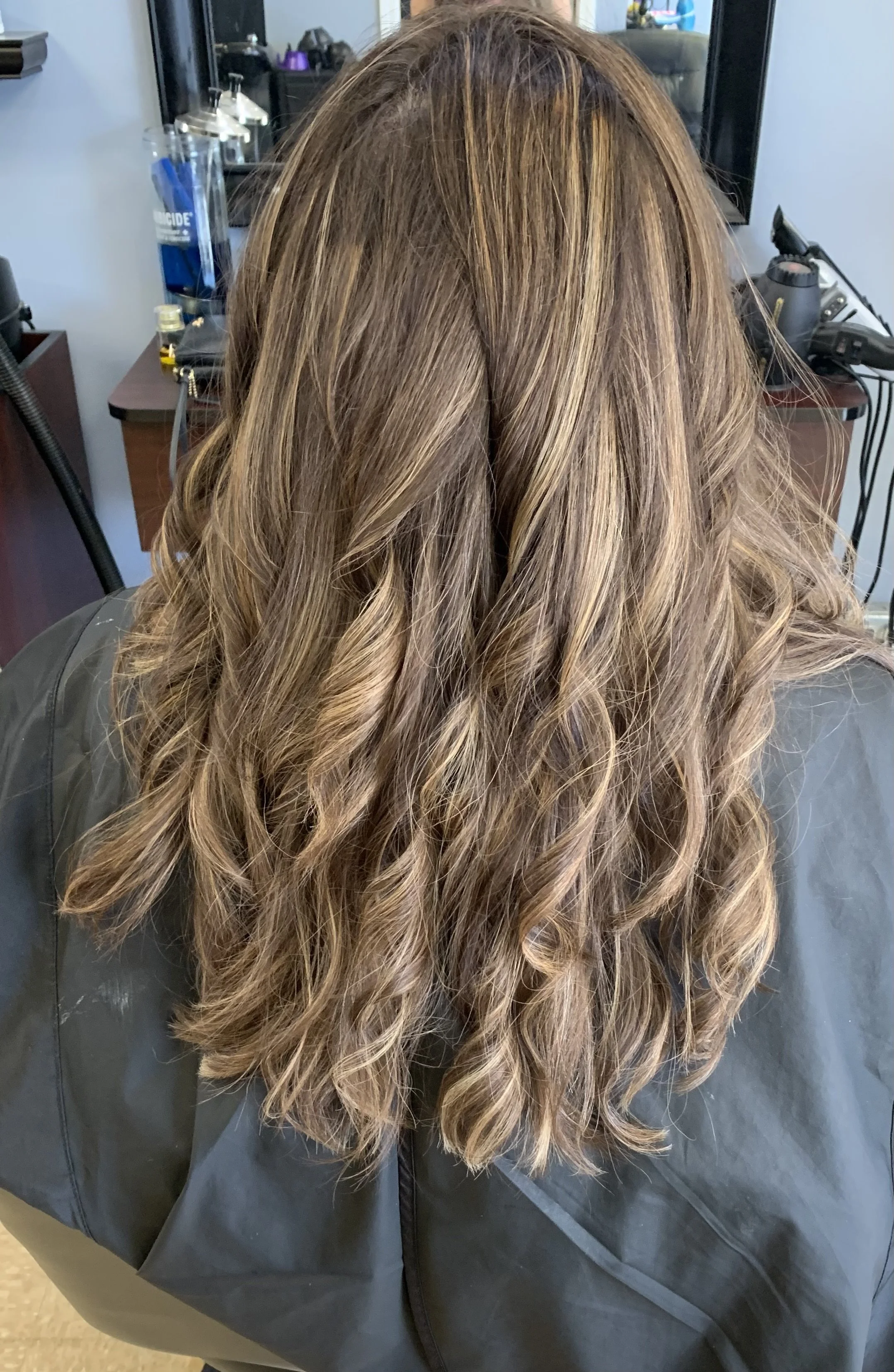 A person with long, wavy, caramel-colored hair sitting with her back to the camera in a salon, wearing a dark cape.