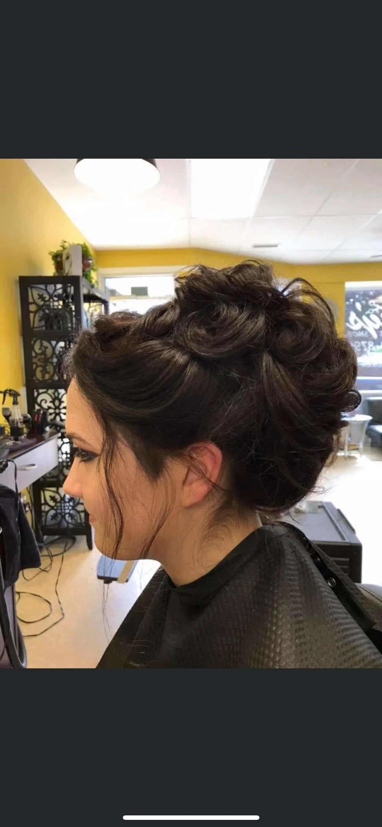 Side view of a woman with styled brunette hair in a salon with yellow walls, styling tools on a black shelf, and a large window in the background.