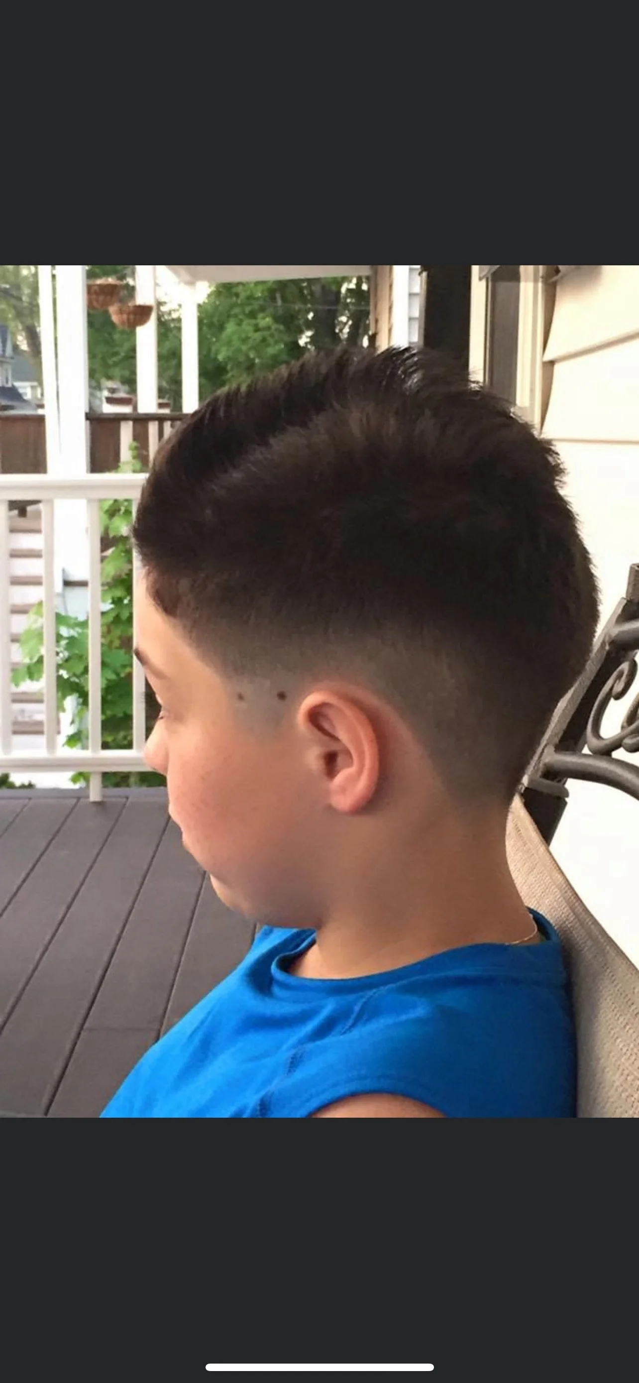 Side profile of a young boy with a fresh fade haircut, sitting on a porch with a wooden deck, white railing, and greenery in the background.