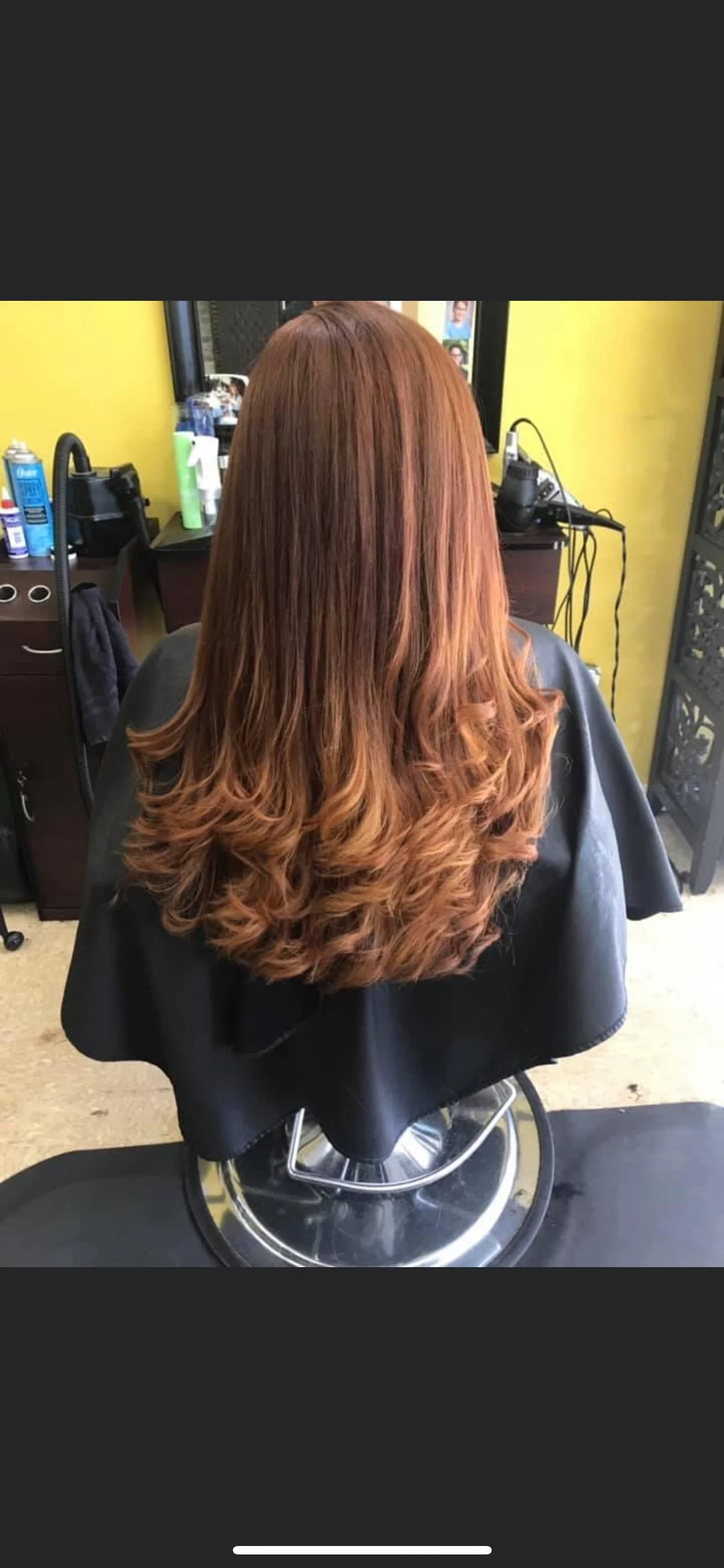 A woman with long, styled, reddish-brown hair sitting in a salon chair with a black cape.
