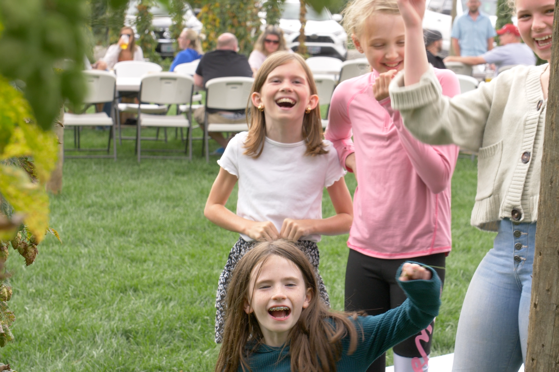Group of children playing outside in a park or garden, smiling and laughing, with some adults and tables in the background.