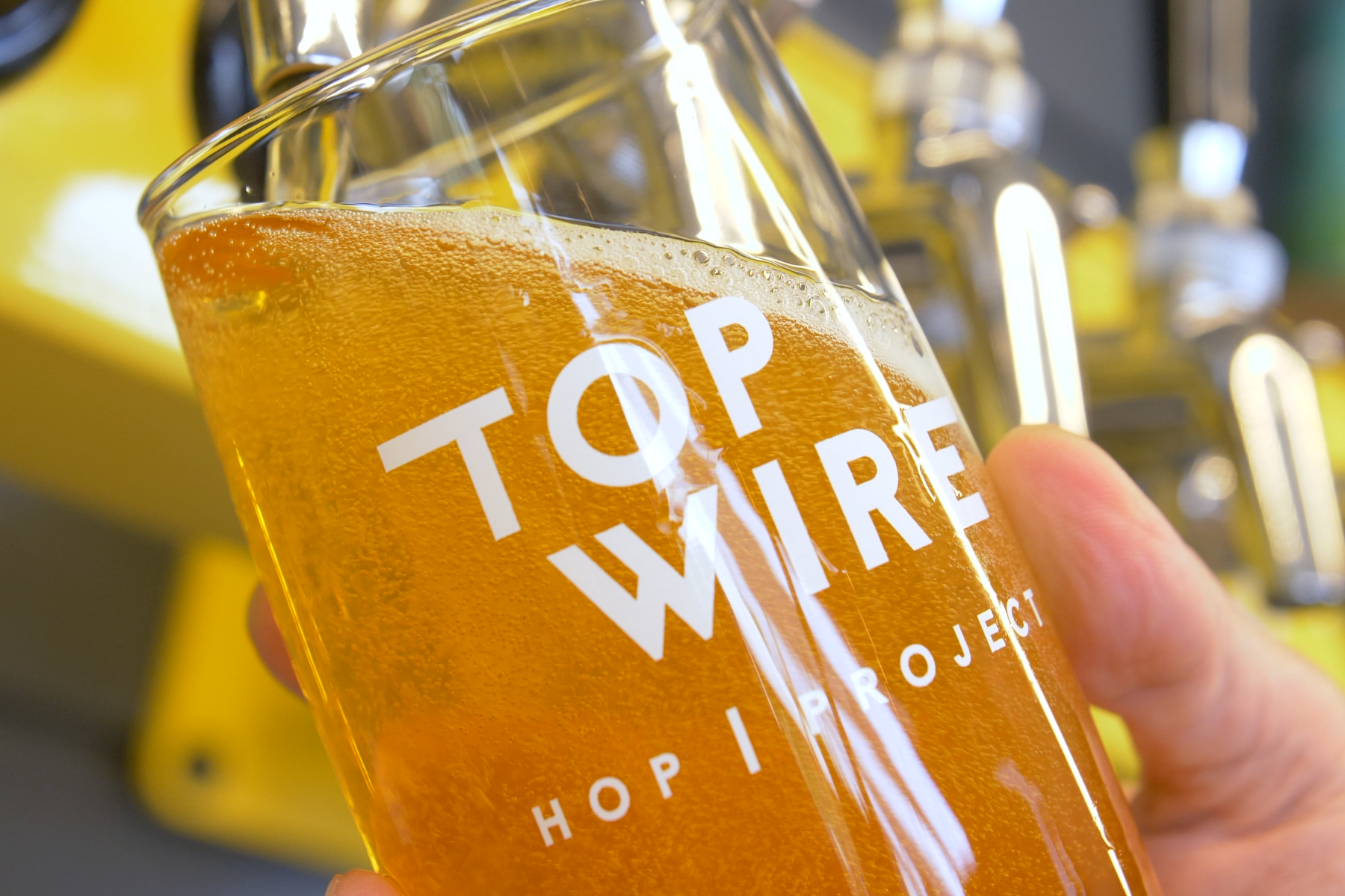 A close-up of a glass of beer being held by a person's hand, with the words 'Top WIRE Hop Project' printed on the glass. The background shows beer taps and yellow accents.