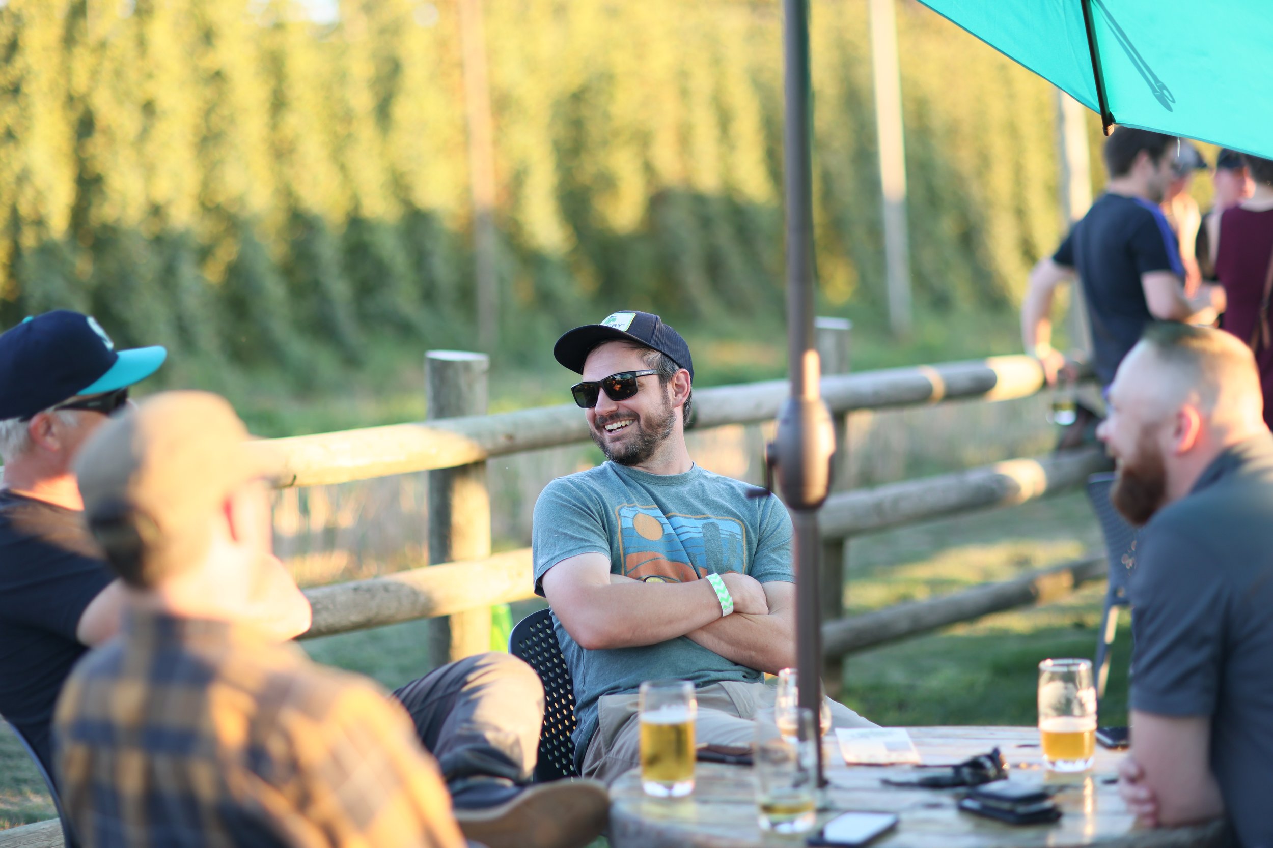 Group of people enjoying outdoors at a social gathering, sitting at a wooden table with drinks, wearing casual clothing, outdoors on a sunny day.