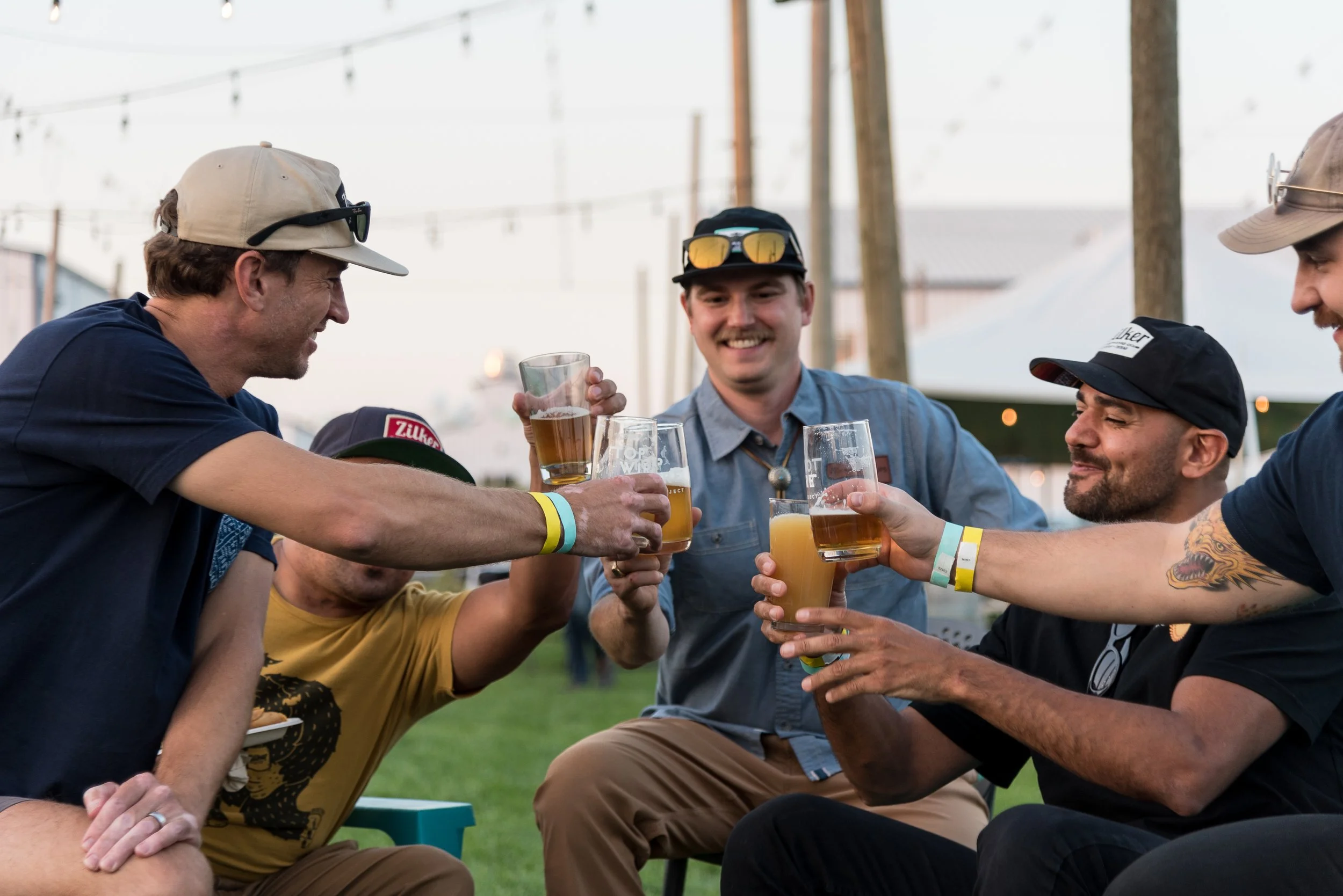 Group of five men sitting outdoors, sharing drinks and smiling, during a casual event with string lights overhead.
