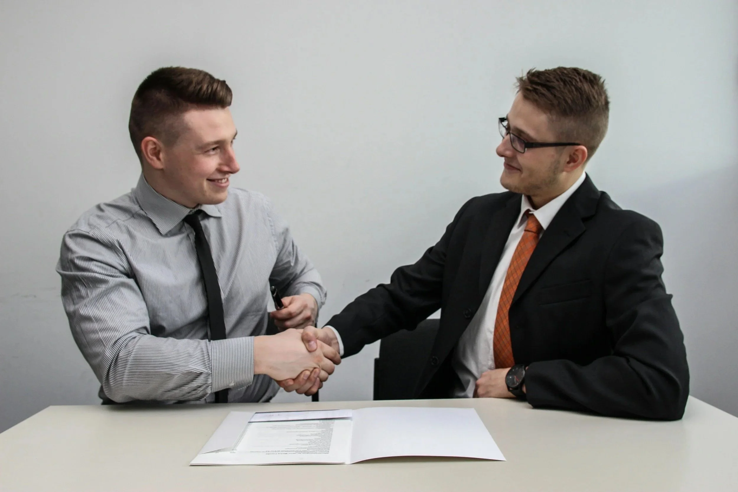 Two men in business attire shaking hands across a table with an open document, smiling at each other.