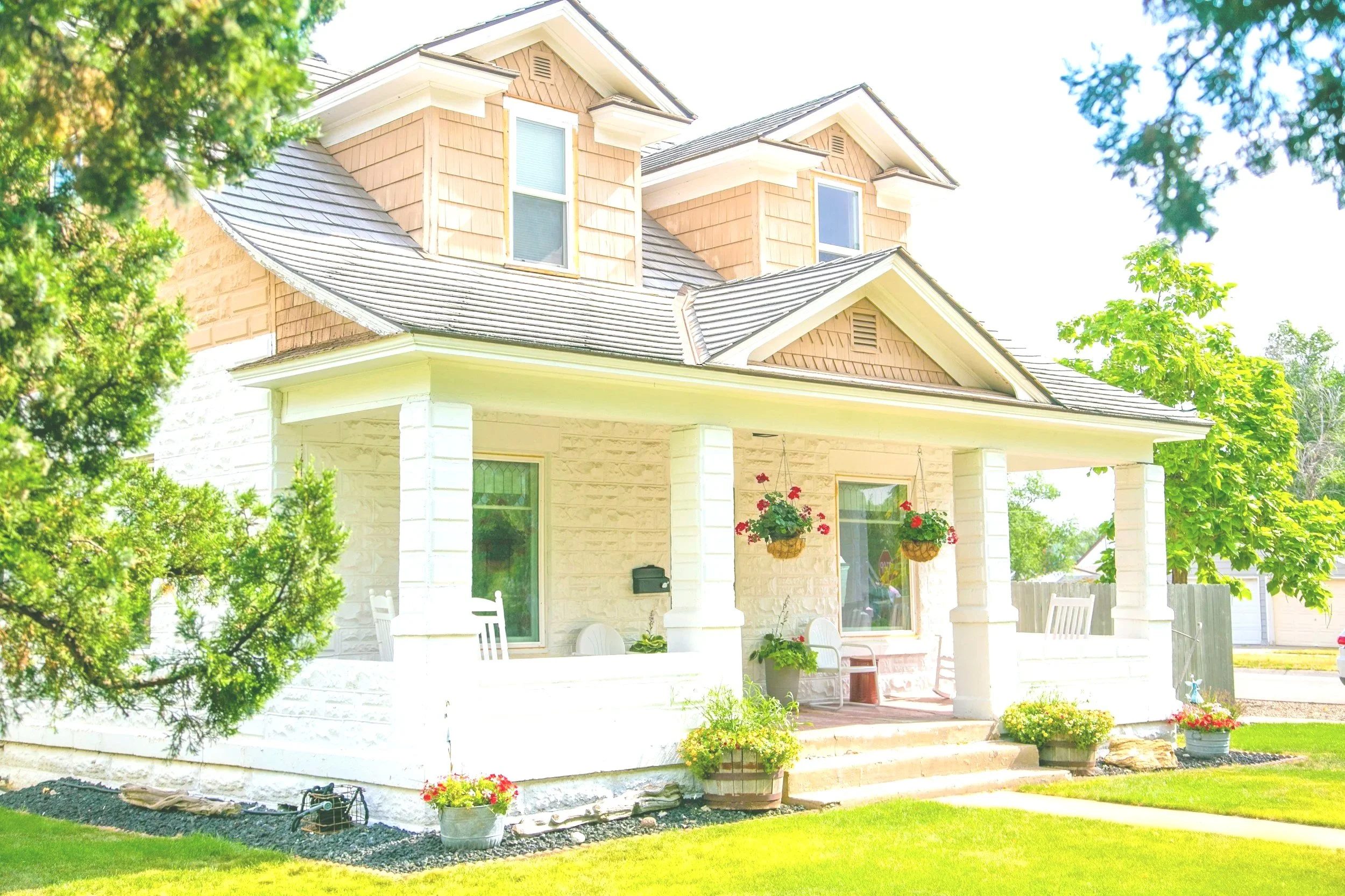 Front view of a two-story house with a porch, beige siding, white stone foundation, hanging flower baskets, potted plants, and a well-maintained lawn.