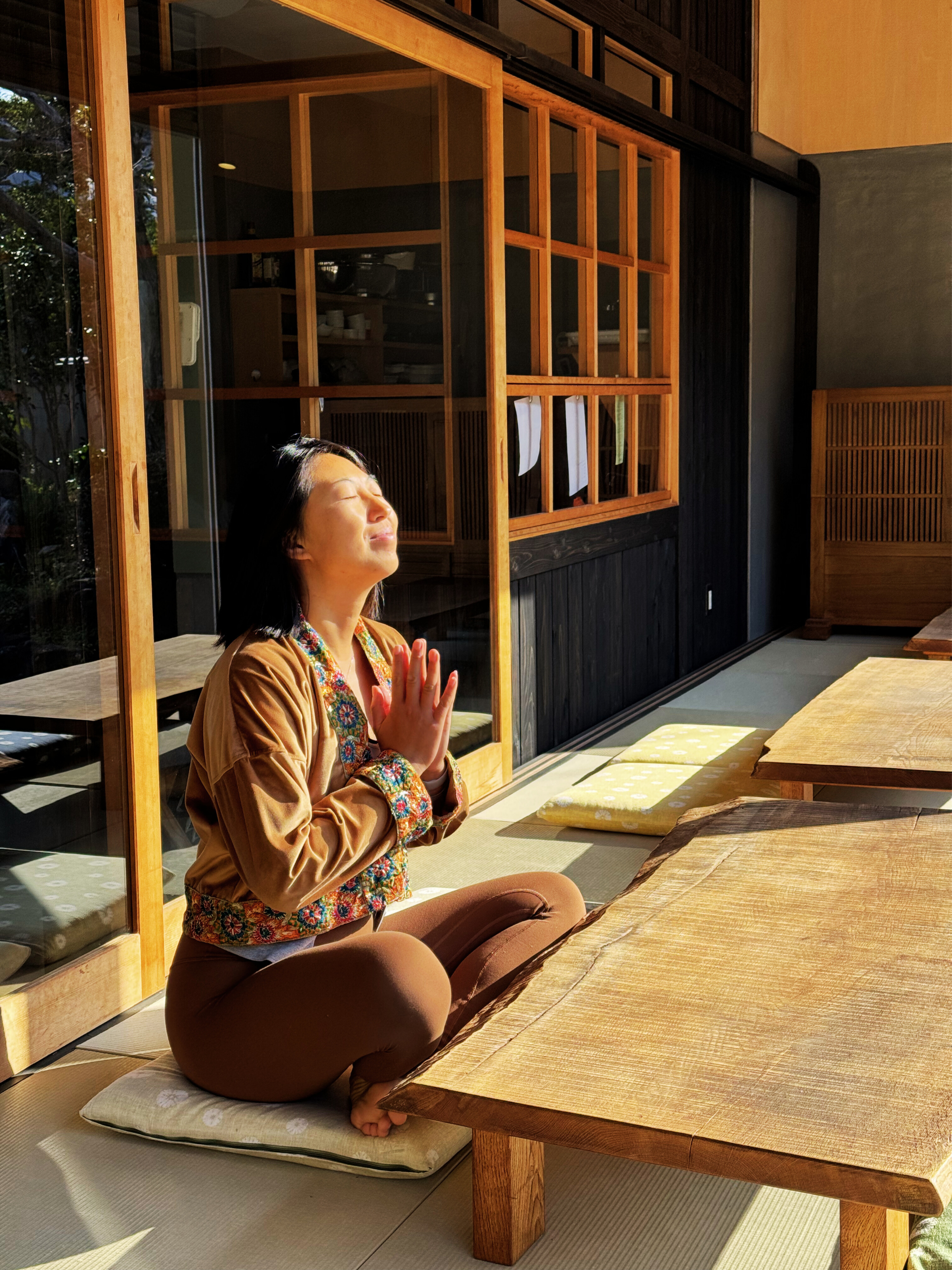 Woman meditating with closed eyes and hands in prayer position near sunlight inside a room with wooden furniture and tatami mats.