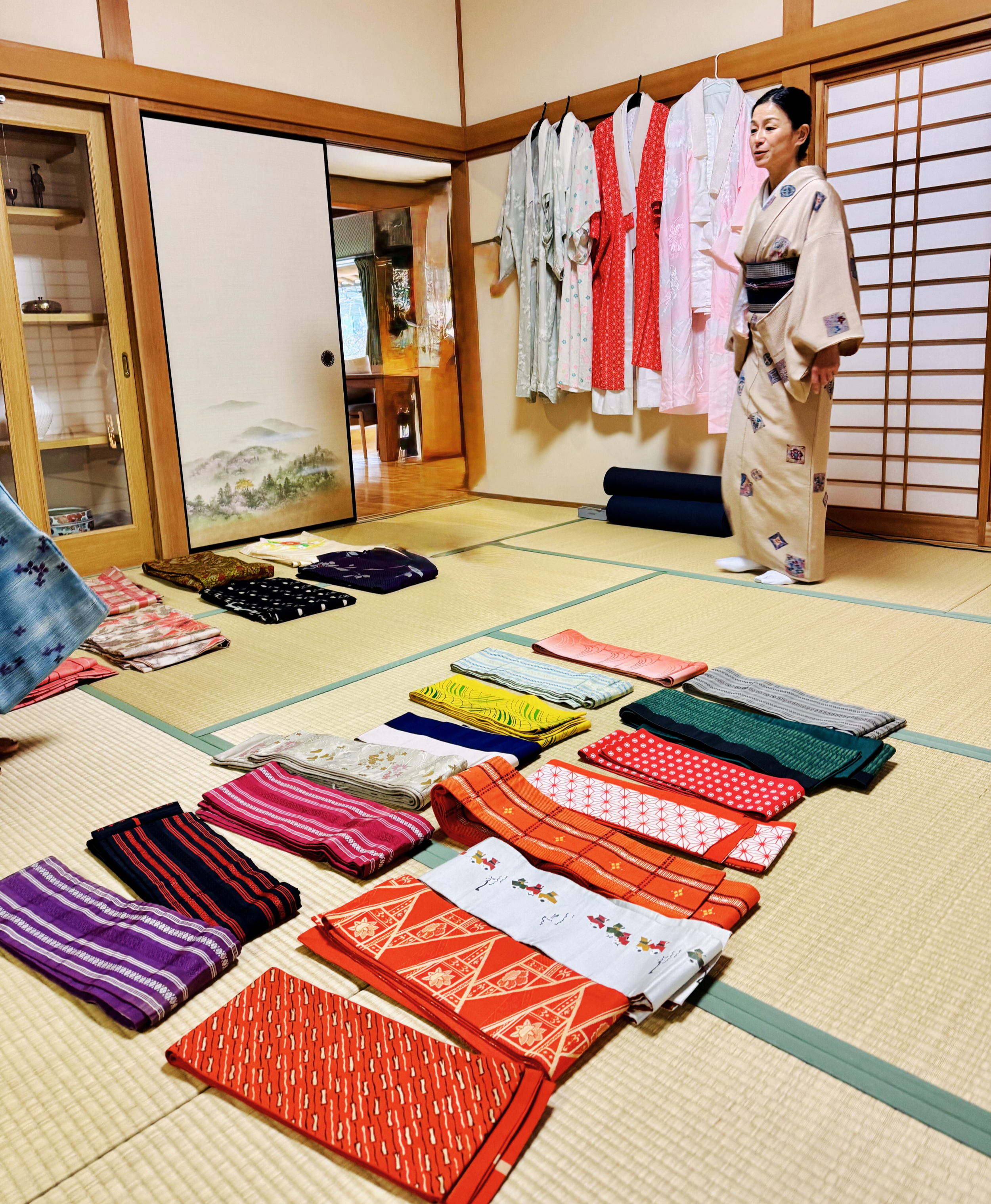 A woman in a kimono standing in a traditional Japanese room with tatami mats, displaying various colorful kimonos and obi sashes laid out on the floor, with additional kimonos hanging on racks on the wall.