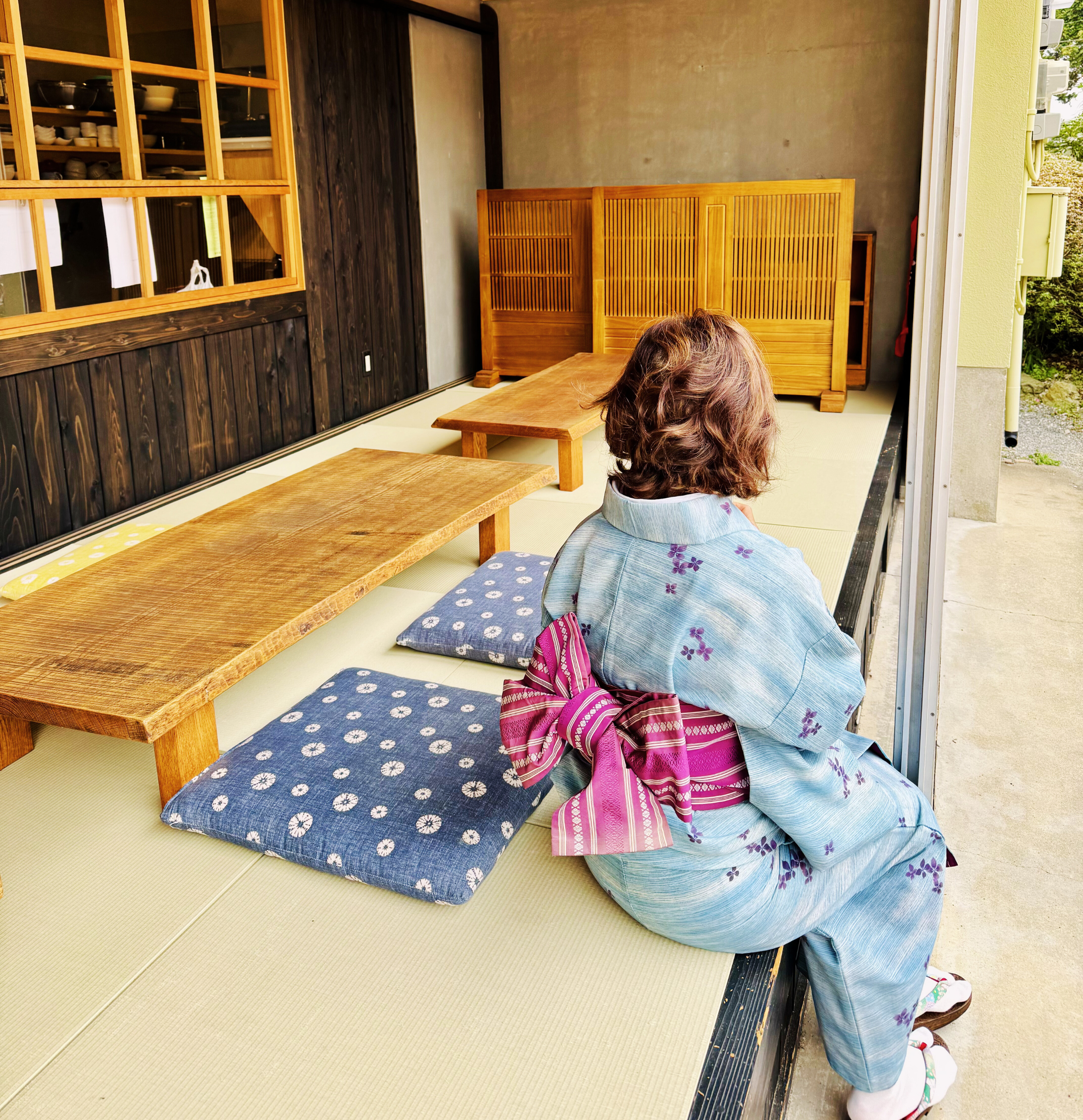 A woman in a traditional Japanese yukata sitting on tatami mats, facing outside, with cushions and low wooden tables around her, in a minimalist Japanese-style room.