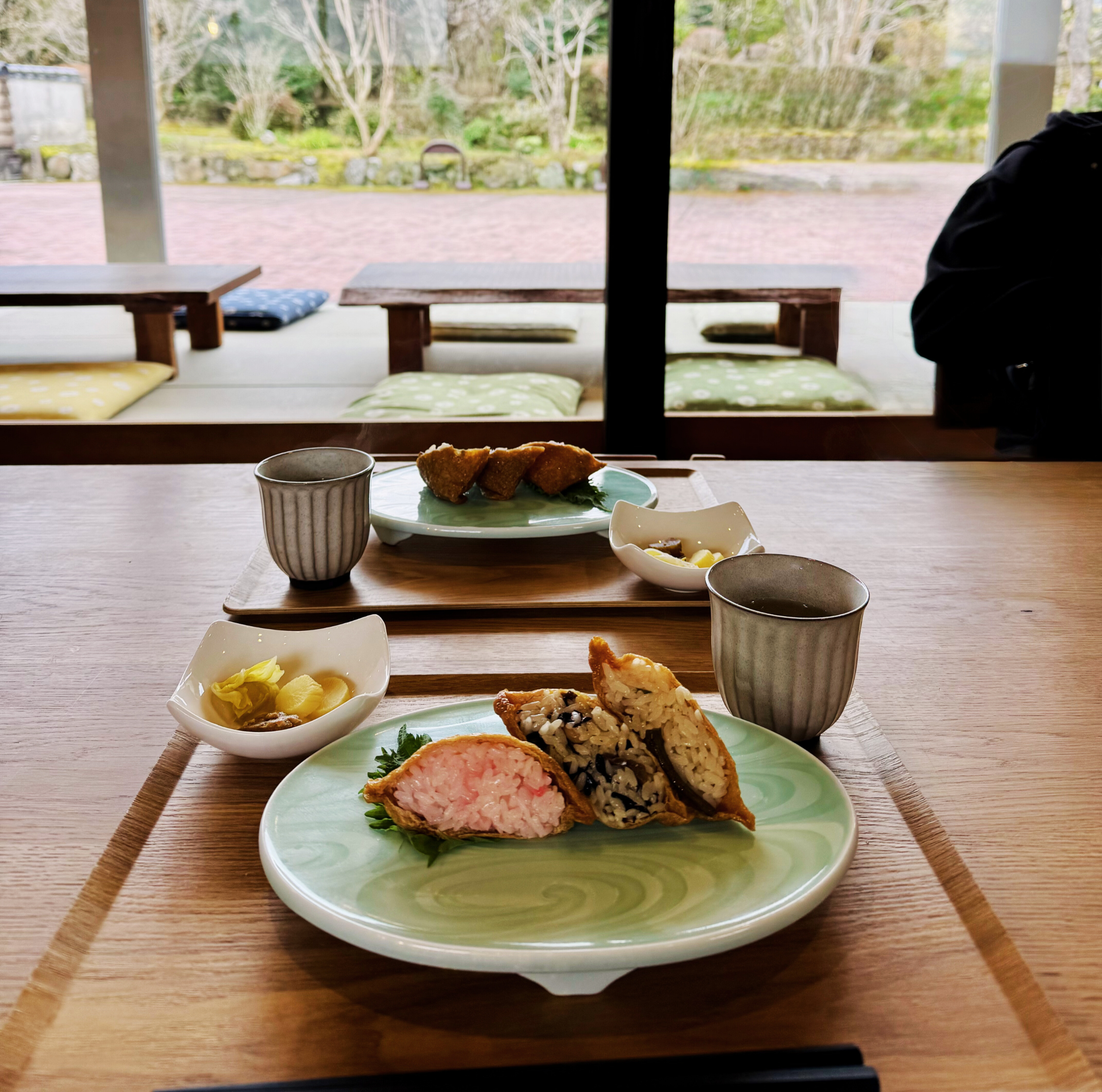 A traditional Japanese meal served on a wooden table with a view of a garden outside. The meal includes rice-filled fried spring rolls, pickled vegetables, and a cup of tea.