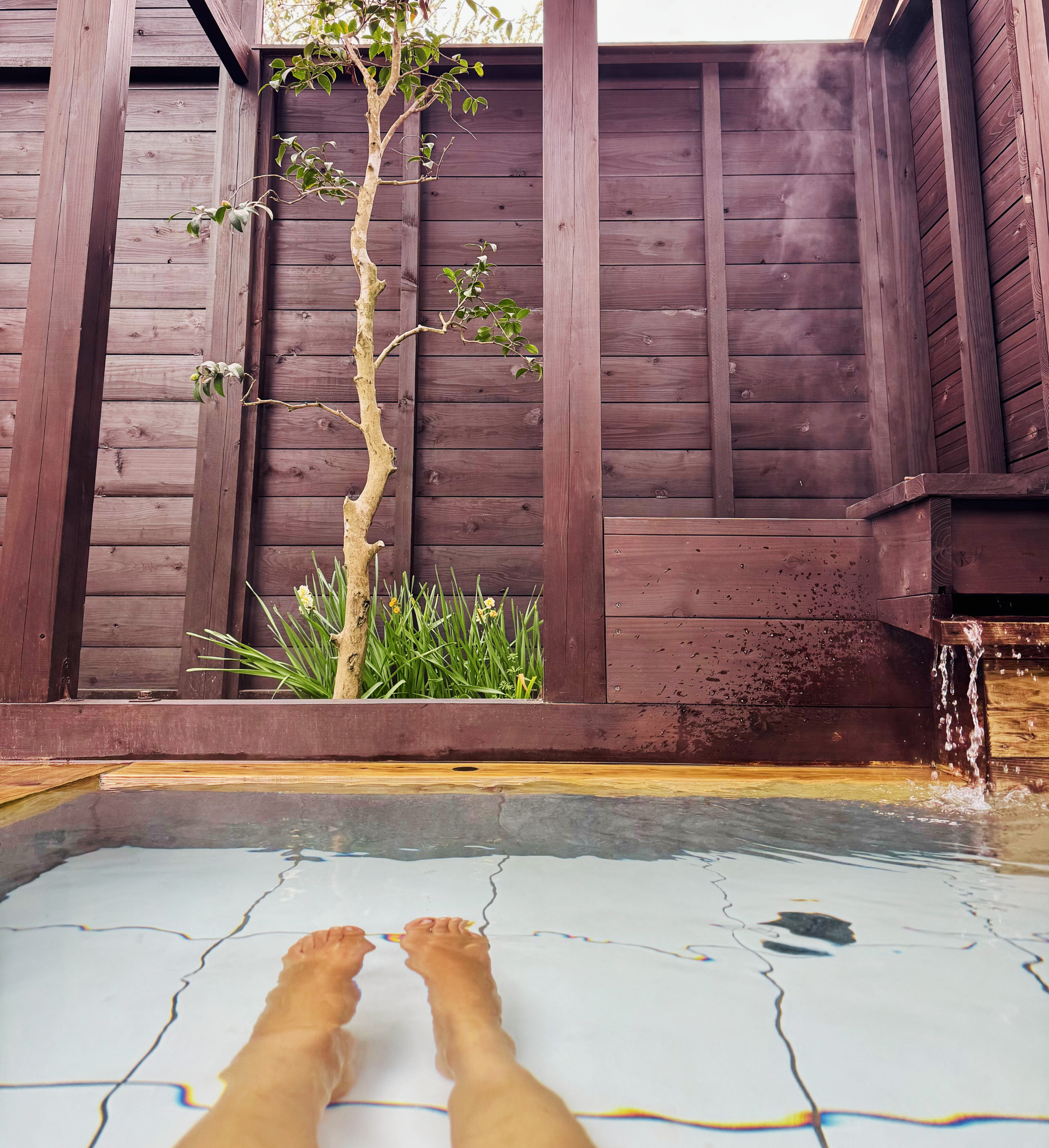 Person's feet submerged in a hot tub next to a wooden privacy fence with a small tree and plants, and a water spout overhead.