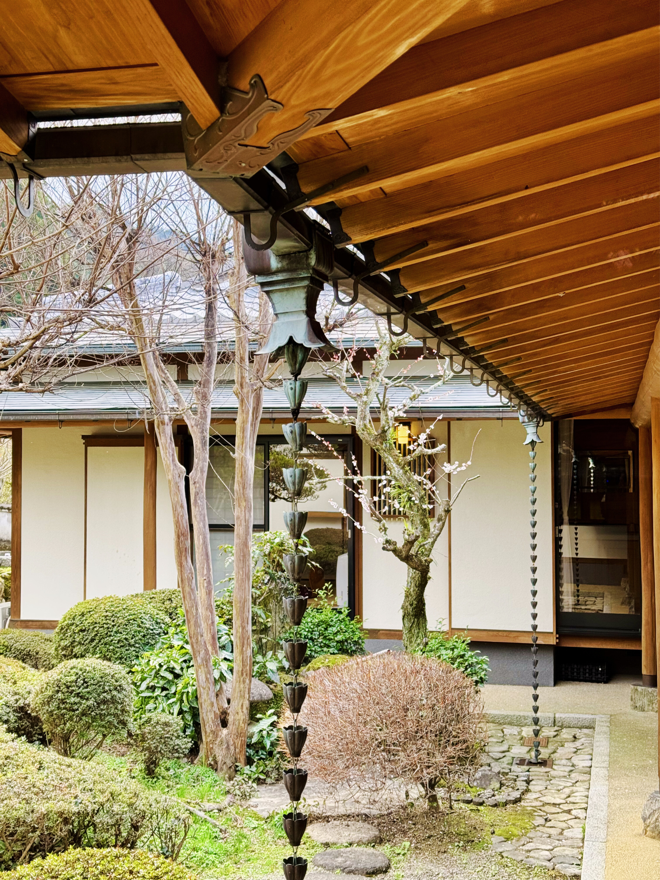 Japanese-style garden with pruning trees, shrubs, and hanging metal wind chimes under a wooden eave.