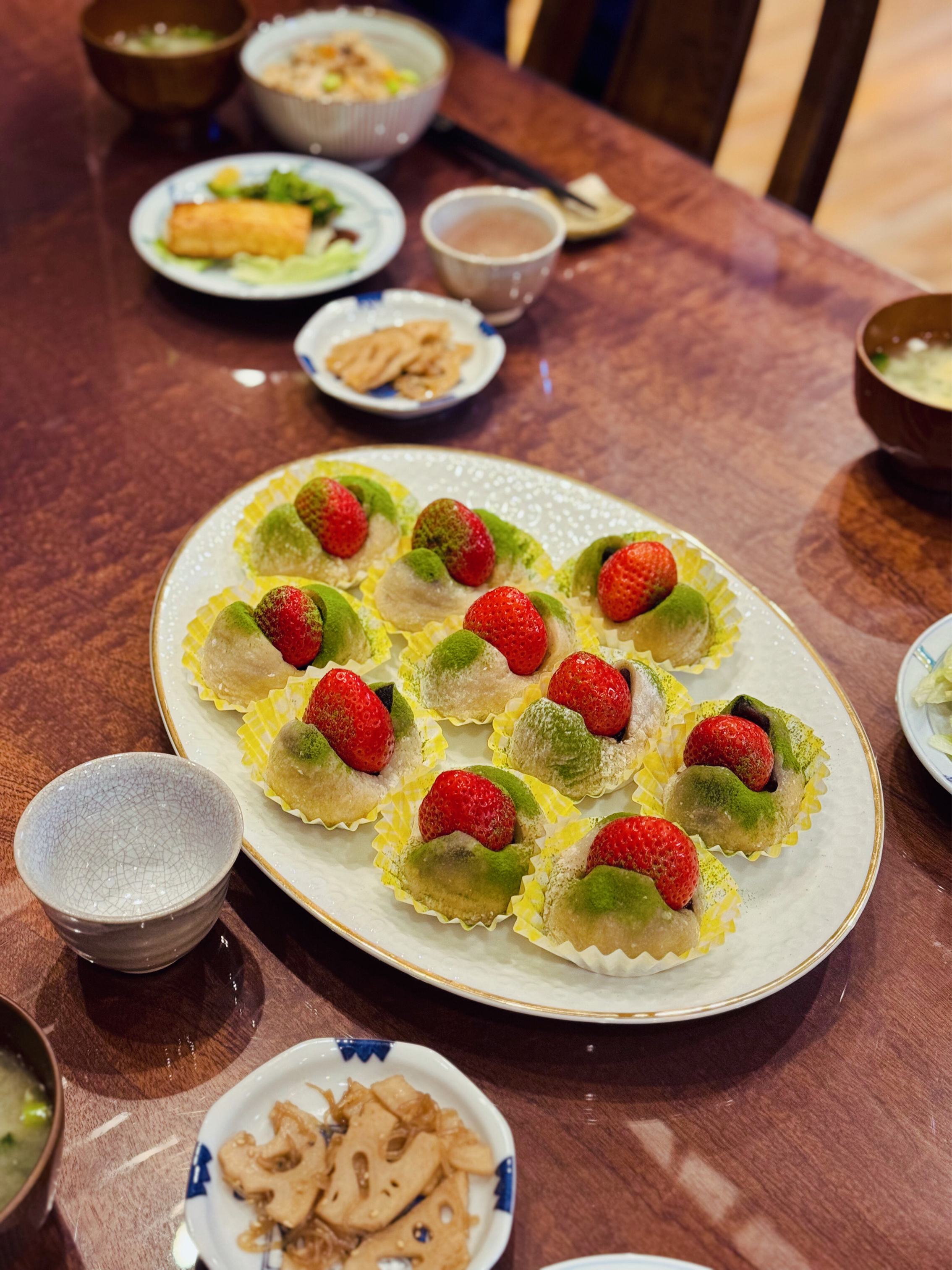 Plate of mochi ice cream topped with strawberries and green matcha powder, surrounded by smaller bowls of assorted Japanese dishes including pickled vegetables, simmered lotus root, miso soup, and other side dishes on a wooden table.