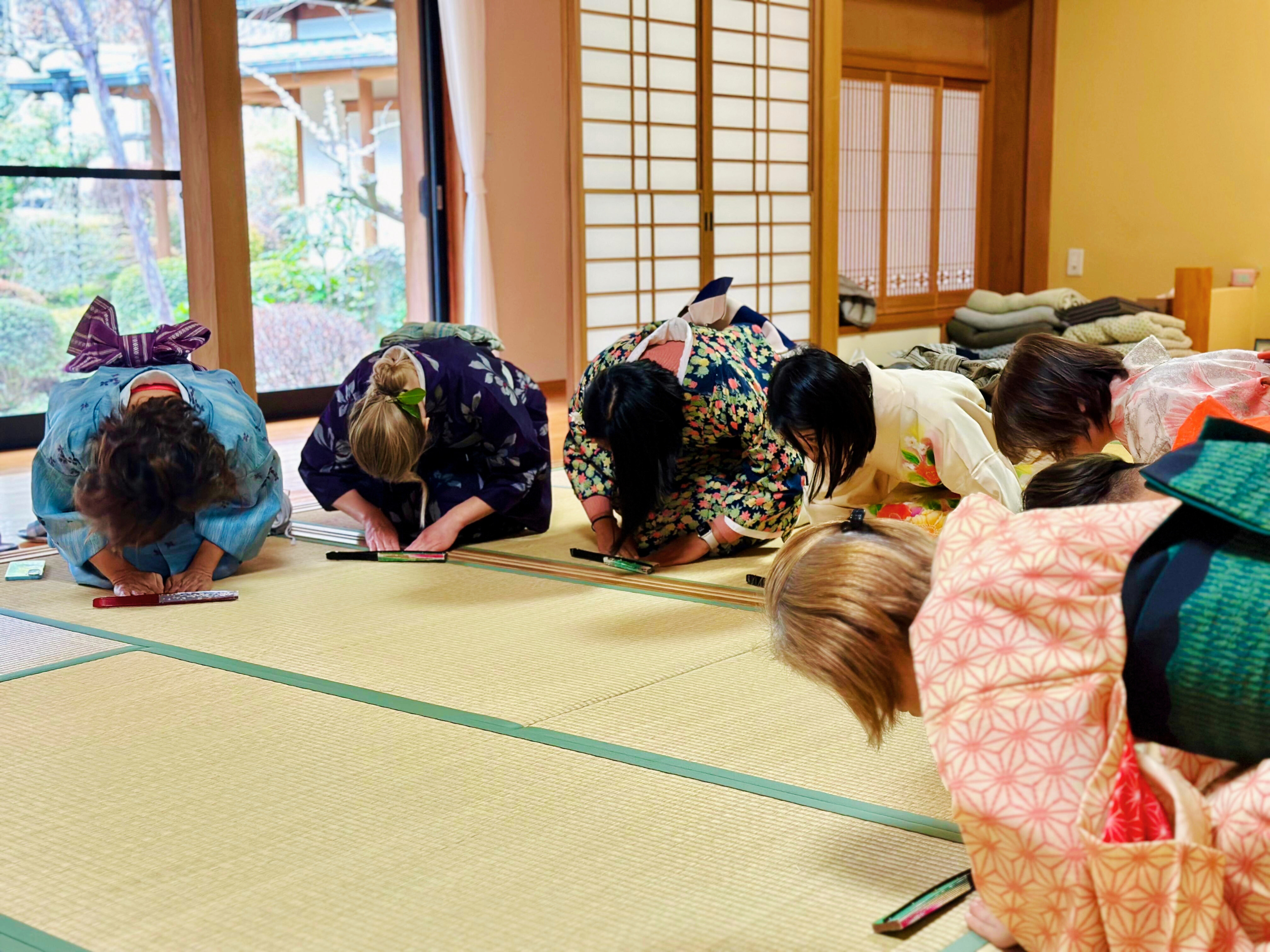 Group of women dressed in colorful kimonos bowing in a traditional Japanese room with tatami mats, sliding shoji doors, and a view of a garden outside.