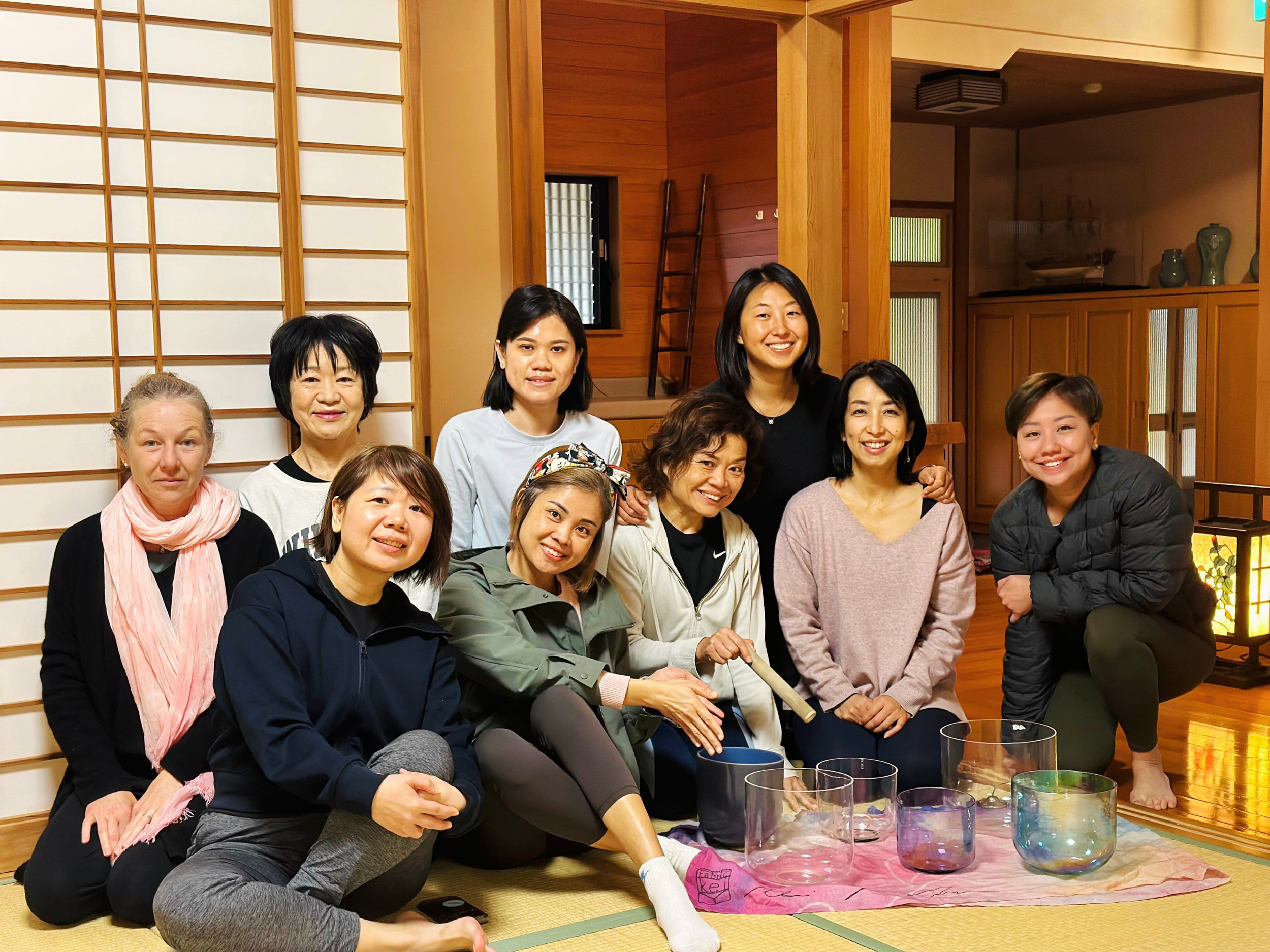 Group of ten women posing together in a traditional Japanese room with wood and shoji sliding doors, some sitting on the tatami floor and others kneeling or squatting behind, smiling for the camera, with crystal singing bowls in front of them.