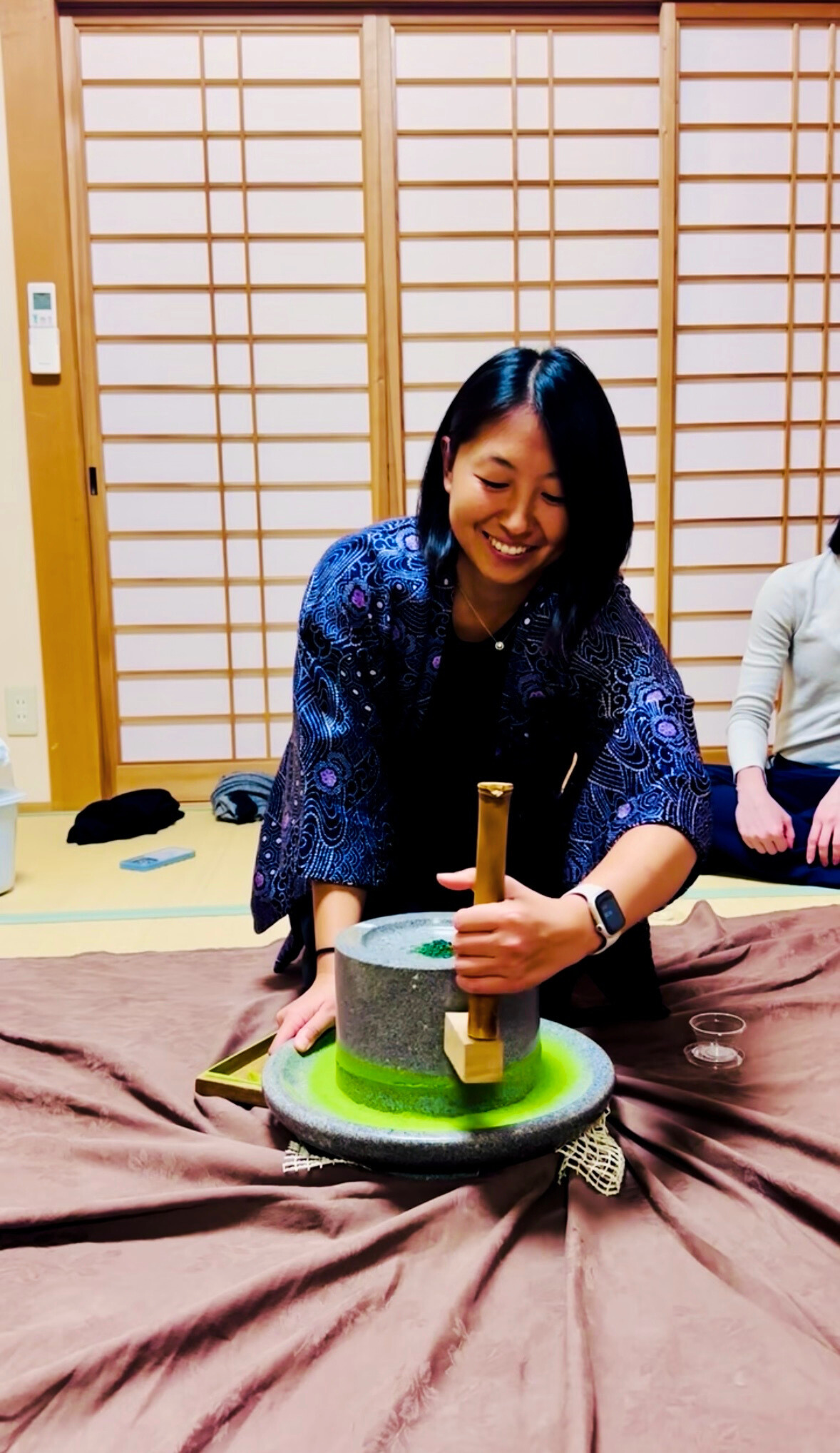 A woman using a traditional Japanese stone mill to grind green tea leaves, with a woman sitting nearby in a room with shoji sliding doors.