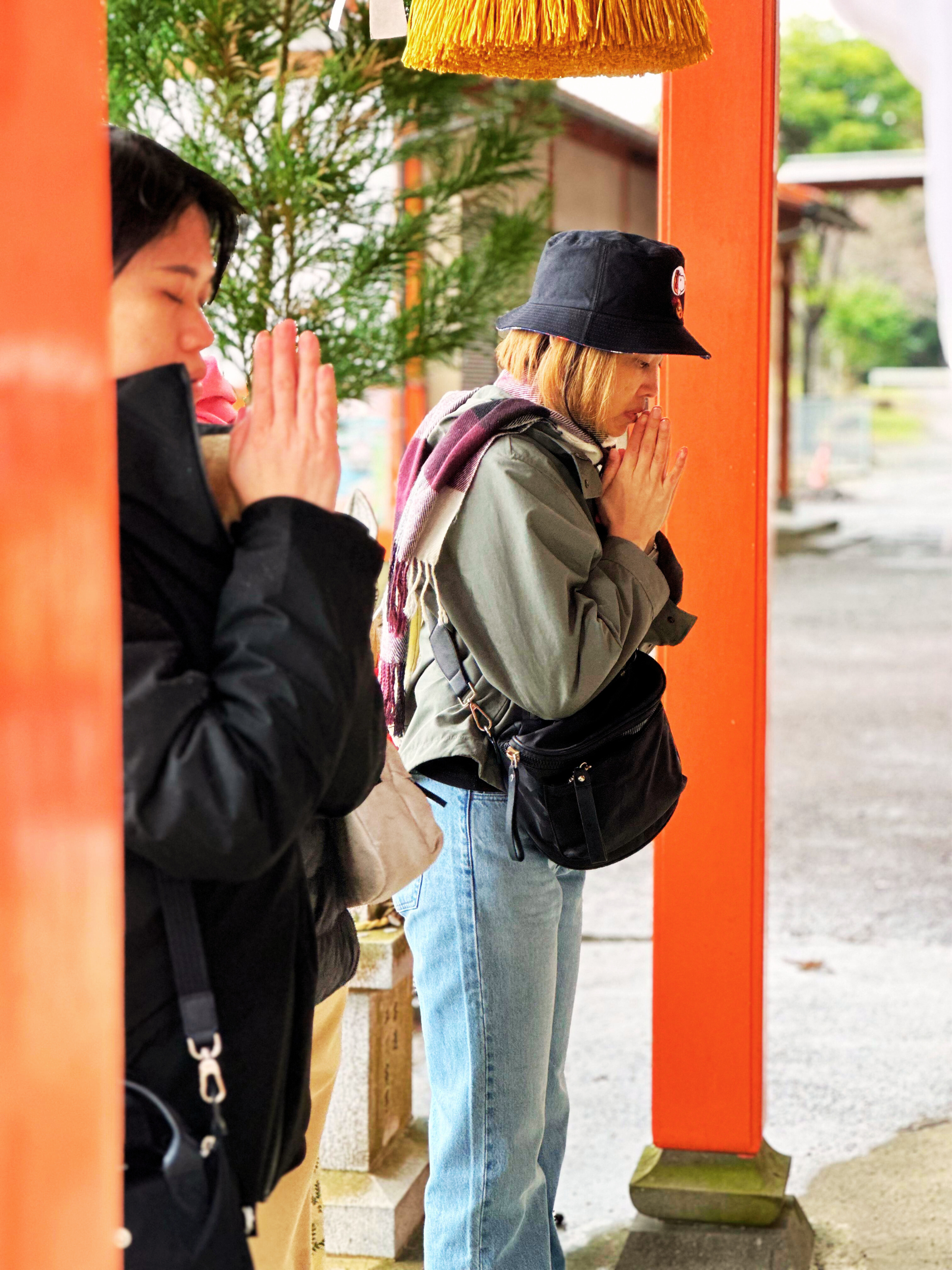 Three women standing outdoors at a shrine, praying with hands pressed together, eyes closed, and heads bowed. They are dressed in casual clothing and have their eyes closed in prayer.