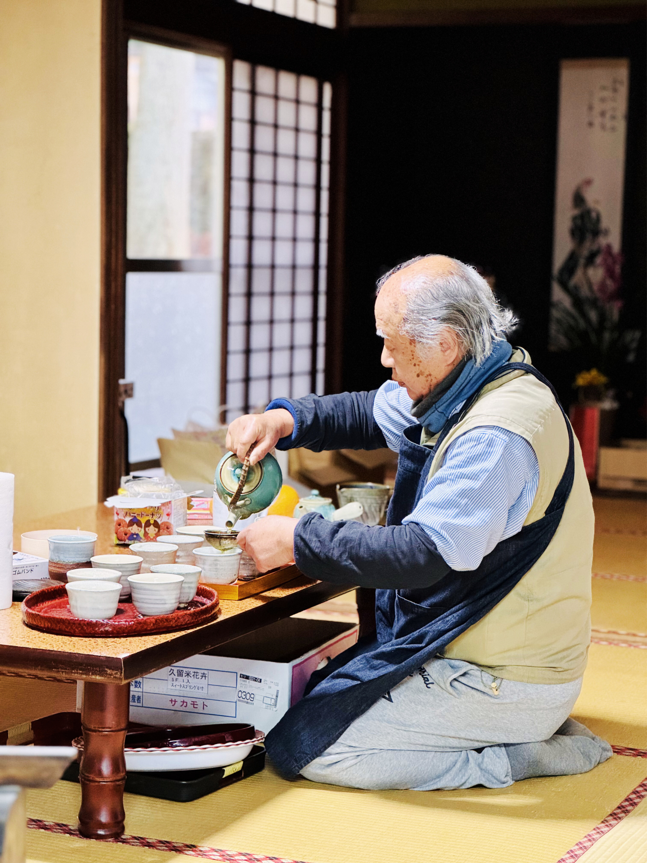 An elderly man with grey hair kneeling on the floor, pouring tea into small cups on a table in a traditional Japanese room.