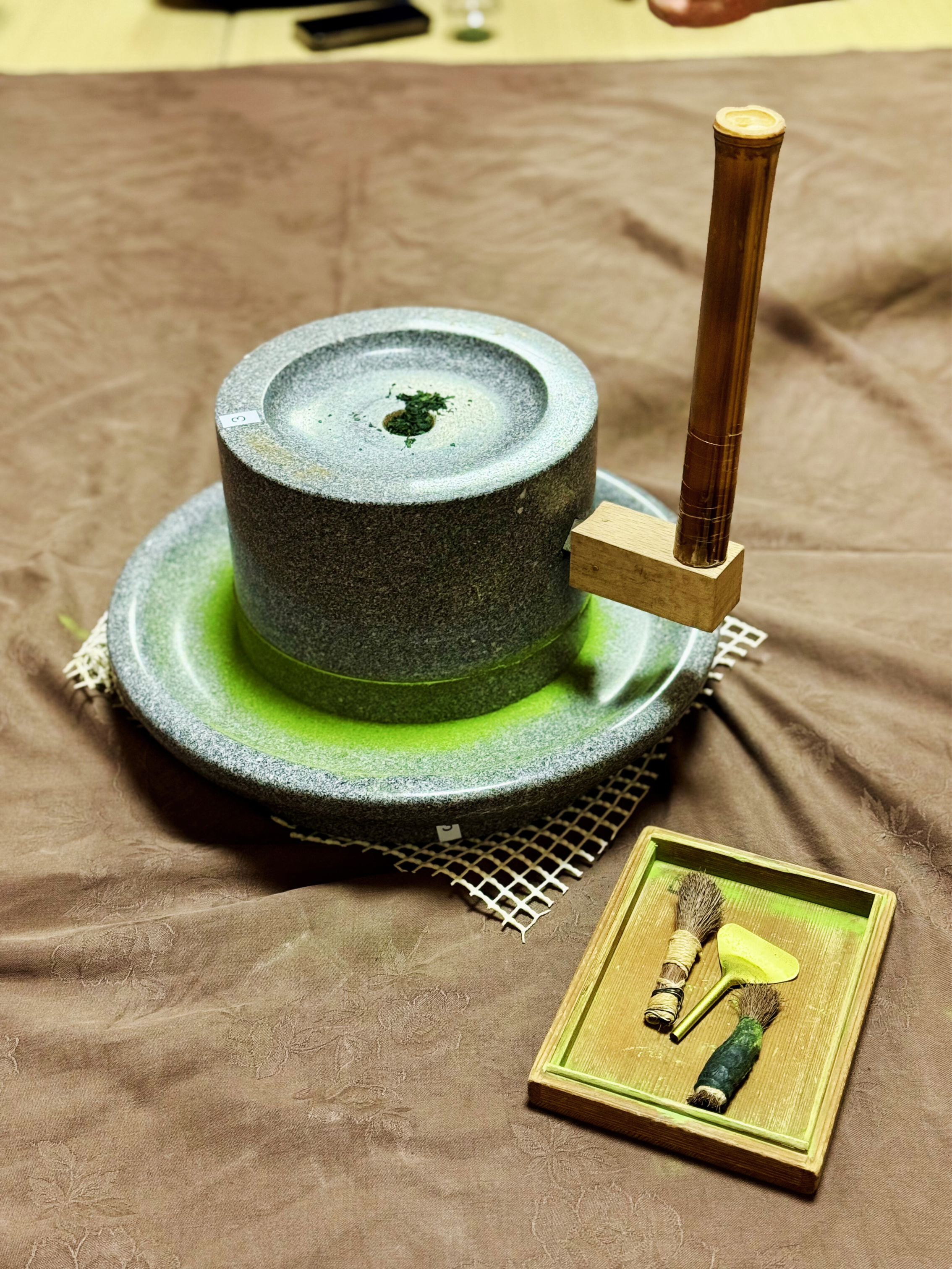 Traditional stone mortar and pestle used for pounding herbs or spices, with a wooden pestle resting on a block, and a small tray holding two small brushes and a yellow spatula.