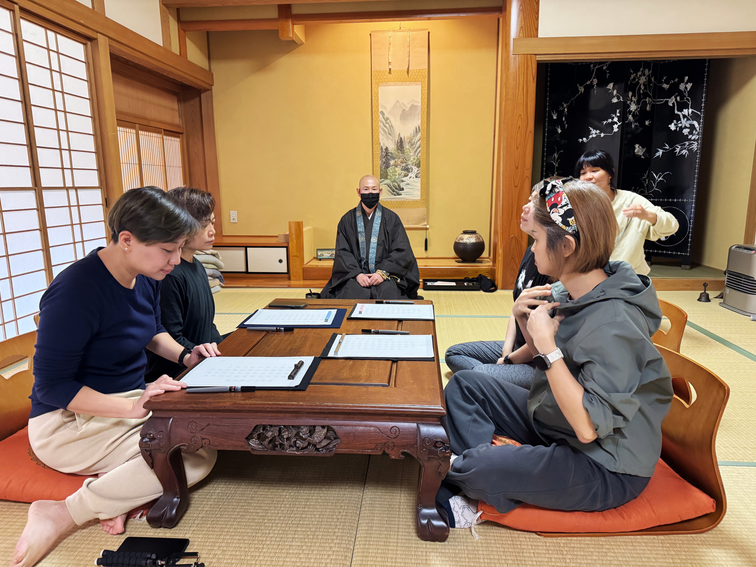 Group of people sitting on floor cushions around a low wooden table inside a traditional Japanese room with tatami mats and sliding shoji doors, engaged in discussion or activity.