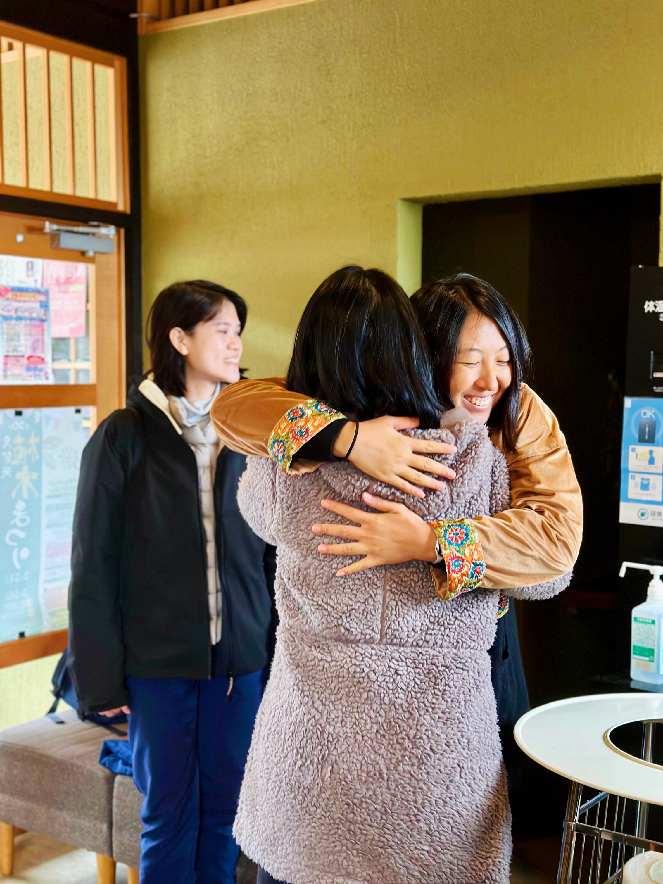 Three women sharing a joyful embrace indoors, with one woman smiling warmly at the center as they hug.