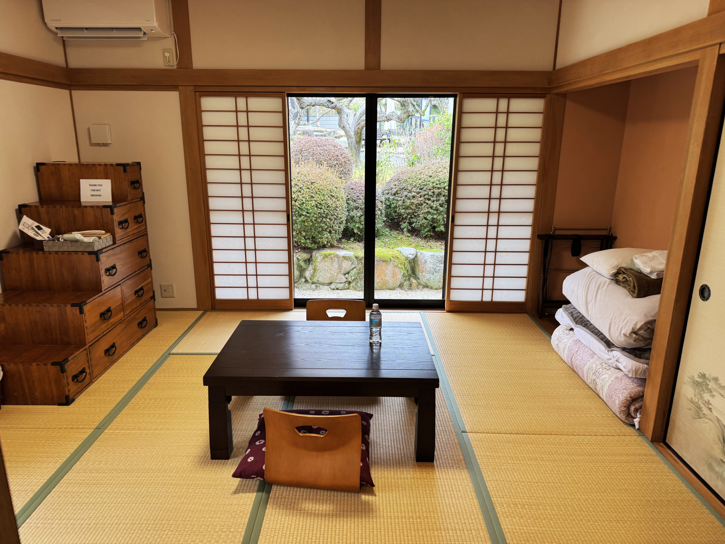 A traditional Japanese tatami room with a view of a garden through sliding shoji doors, containing a low table, cushions, a wooden chest of drawers, and neatly stacked bedding.